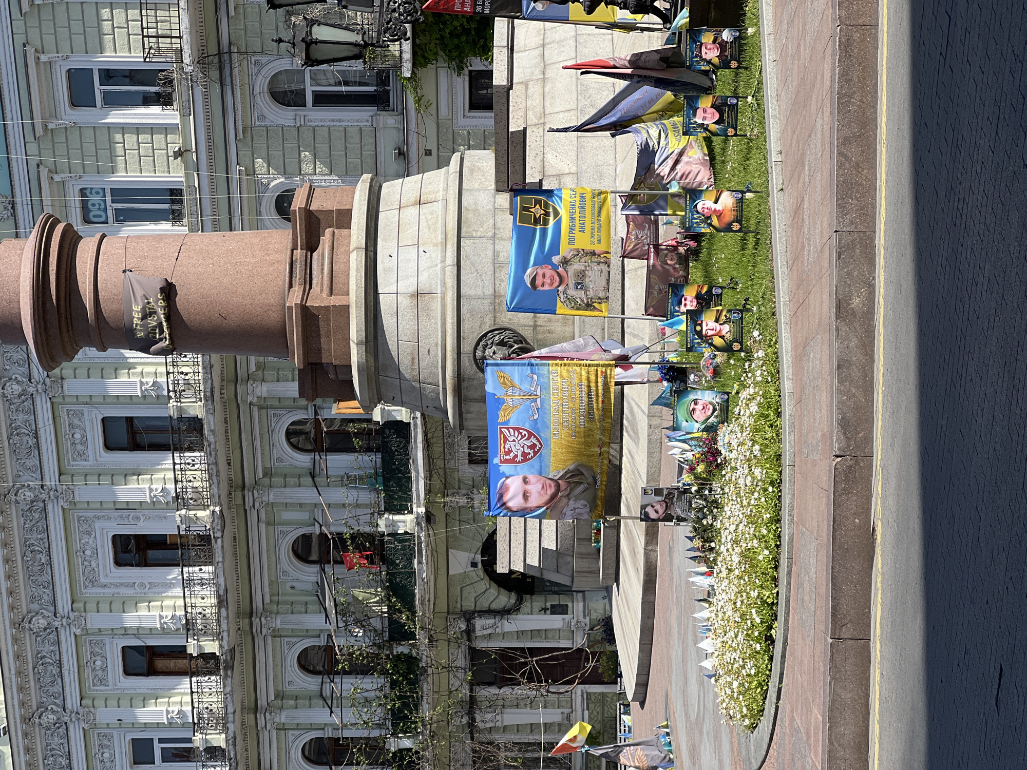 This image shows the Potemkin Steps in Odesa, Ukraine. Memorial banners with portraits of Ukrainian soldiers are displayed around a monument. Some banners feature military symbols and text in Ukrainian. Flowers and small flags are placed as tributes. The architectural facade of a building can be seen in the background. A section of a road is also visible, casting a shadow.