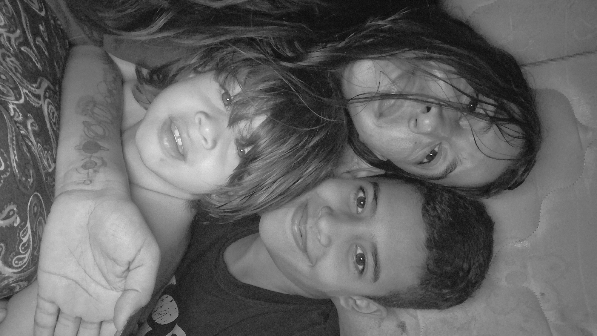 A black and white, close-up photograph depicts an adult female, a teenage boy, and a young child positioned closely together in the city of Vallecito, Venezuela. The adult female, with long, dark, disheveled hair partially obscuring her face, is in the top left, looking towards the camera with a slightly open mouth. To her right, a teenage boy with short, dark hair smiles broadly at the camera. In the foreground, below the other two, a young child with shaggy dark hair also looks towards the camera with a slight smile, revealing front teeth. The child's left forearm, featuring a tattoo or drawing of celestial bodies (planets and rings), is prominent at the bottom of the frame, resting on a dark fabric with a light paisley pattern. The background behind the adult and teenager shows a light-colored, textured surface, possibly a mattress. The subjects are tightly composed, indicating a close relationship and a posed photograph taken indoors.