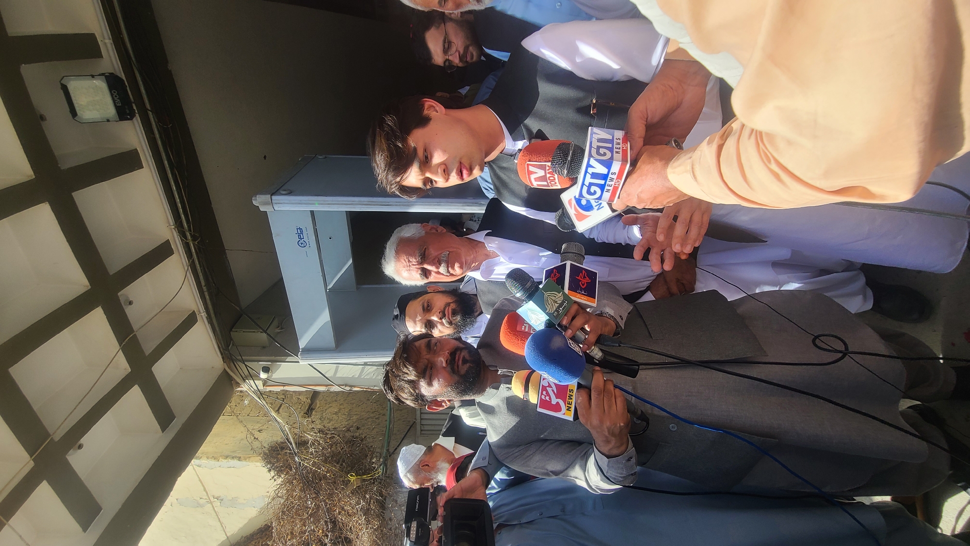 The image shows a group of men surrounded by microphones with various news organization logos on them, suggesting a press conference or interview setting. The men are dressed in a mix of traditional and formal wear. The foreground features several microphones held by unseen individuals, with visible logos of GTV and other news outlets. In the background, there is a glimpse of a building exterior. The image was taken in Quetta City Tehsil, Pakistan.