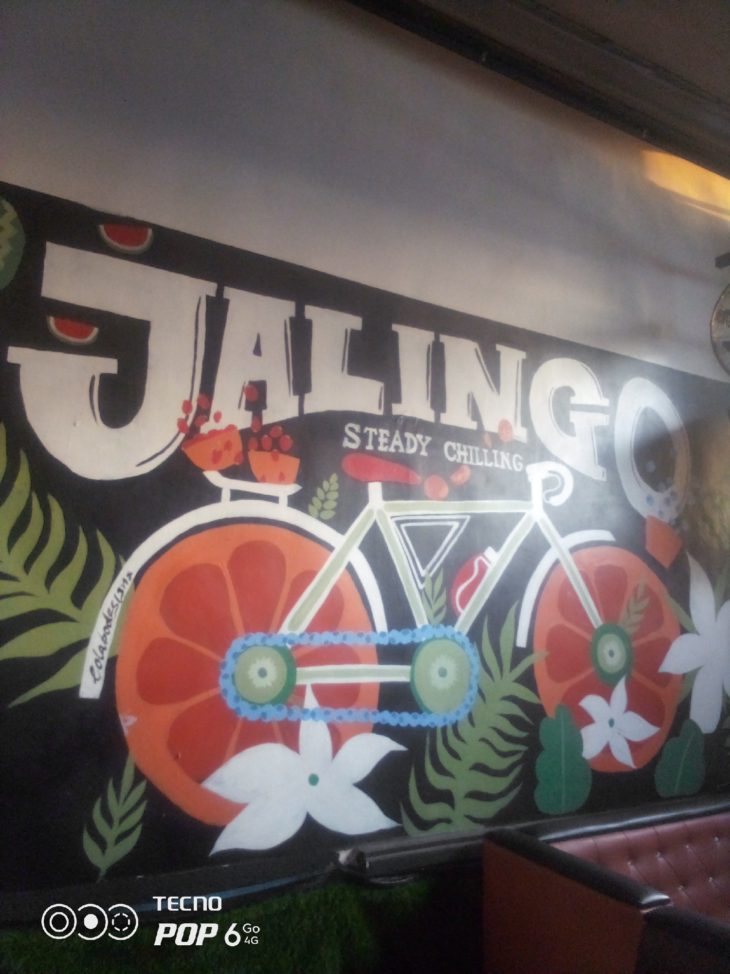 The image displays a mural painted on a dark wall, likely inside a building. The mural features large, white lettering spelling "JALINGO" across the top, with the words "STEADY CHILLING" below. To the left of the lettering are stylized watermelons slices. The central element of the mural is a white bicycle with its wheels depicted as orange slices. The bicycle has a blue chain and green details. Surrounding the bicycle are various green leaf motifs and white flowers. On the left side of the bicycle's front wheel, a white ribbon with black text is visible, appearing to read "Colabodesigns." At the bottom of the image, the "TECNO POP 6 4G" logo is present, indicating a phone's watermark. The scene appears to be in a public or commercial space, possibly a cafe or restaurant, given the presence of a brown leather booth seat in the foreground. The location is identified as Howayi, Nigeria.
