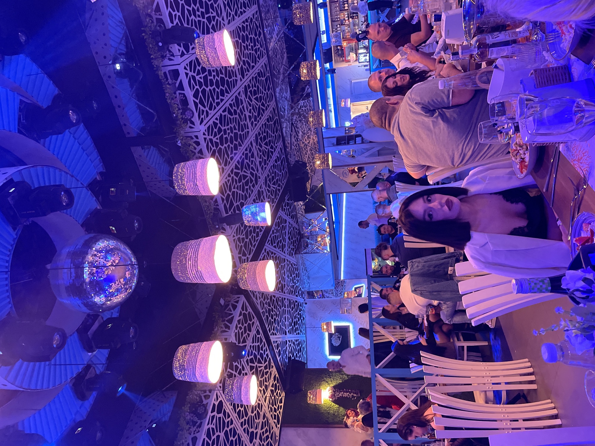 The photograph appears to be taken in a restaurant or bar setting. The location is in Plovdiv, Bulgaria.
The image depicts several individuals seated at tables, engaging in conversation and presumably dining. There is one woman seated nearest the camera. Some people are conversing and dining at the tables. The room is lit with a combination of overhead lighting, including cylindrical hanging lamps, and a disco ball. The ceiling features a complex geometric design. The tables are set with glassware, plates, and bottles. The overall ambiance suggests a lively social atmosphere.