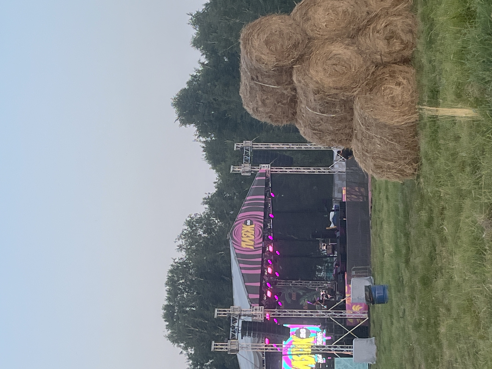 This is a vertical, outdoor photograph taken at twilight in Surakovo, Russia. The scene depicts an outdoor music festival with a stage set up in the center. The stage is adorned with pink and purple lights and features a large screen displaying colorful graphics. The logo "MAXX" is prominently displayed on the stage's canopy. A crowd of people can be seen gathered around the stage, though they are largely obscured by the stage structures. To the right of the stage, a stack of hay bales is piled high, suggesting a rural or agricultural setting. The grass surrounding the stage and hay bales is green and appears to be well-maintained. The sky is a pale blue, indicating the day is coming to an end. The overall atmosphere appears festive and energetic, with the vibrant lights and gathered crowd suggesting an enjoyable musical performance.