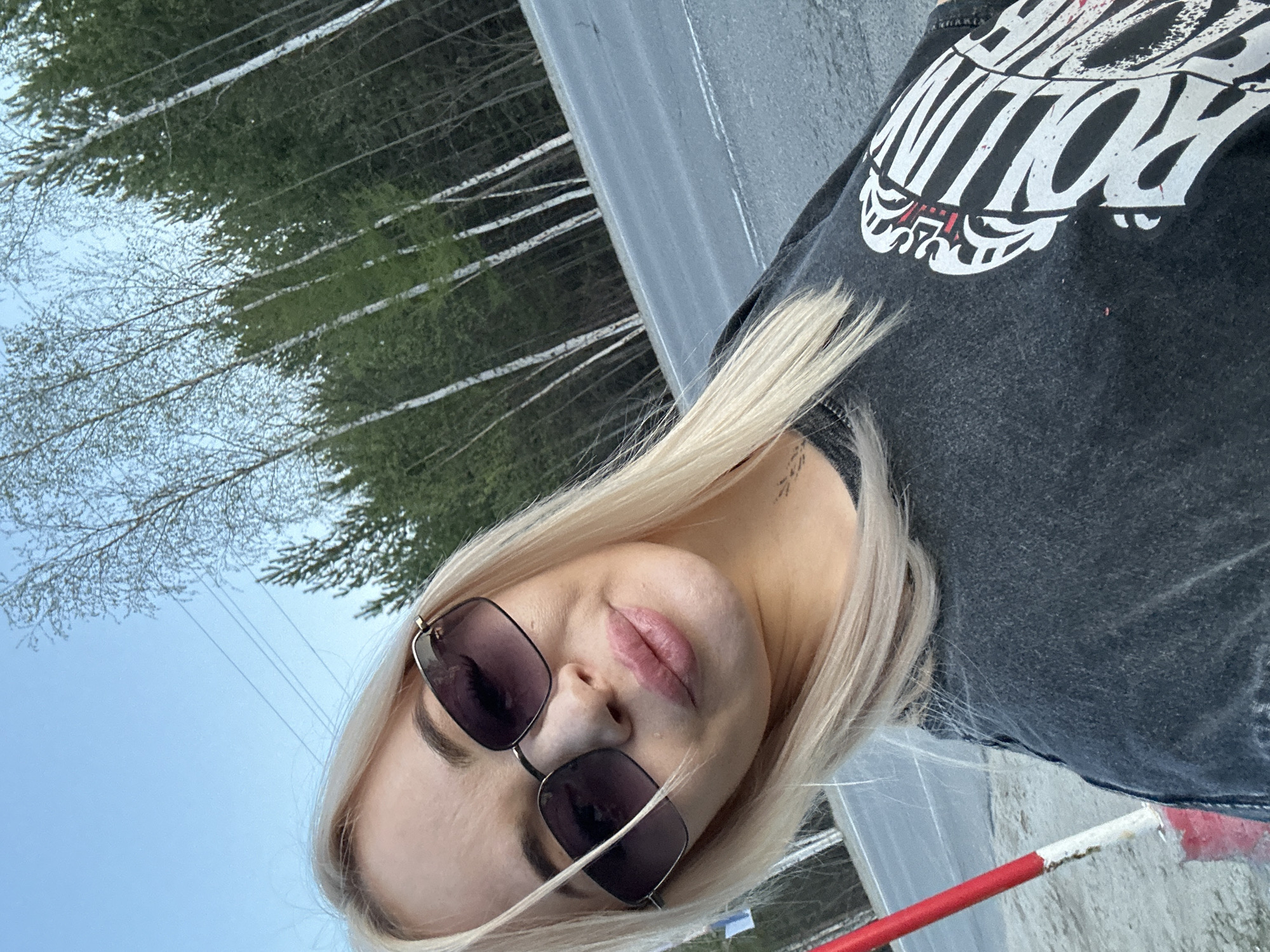 A woman with blonde hair is wearing sunglasses and a dark t-shirt with a white print. She appears to be outdoors, as indicated by the trees in the background, a roadside barrier, and the sky. The location is potentially near the city of Высокий, Russia, as indicated by the query.
