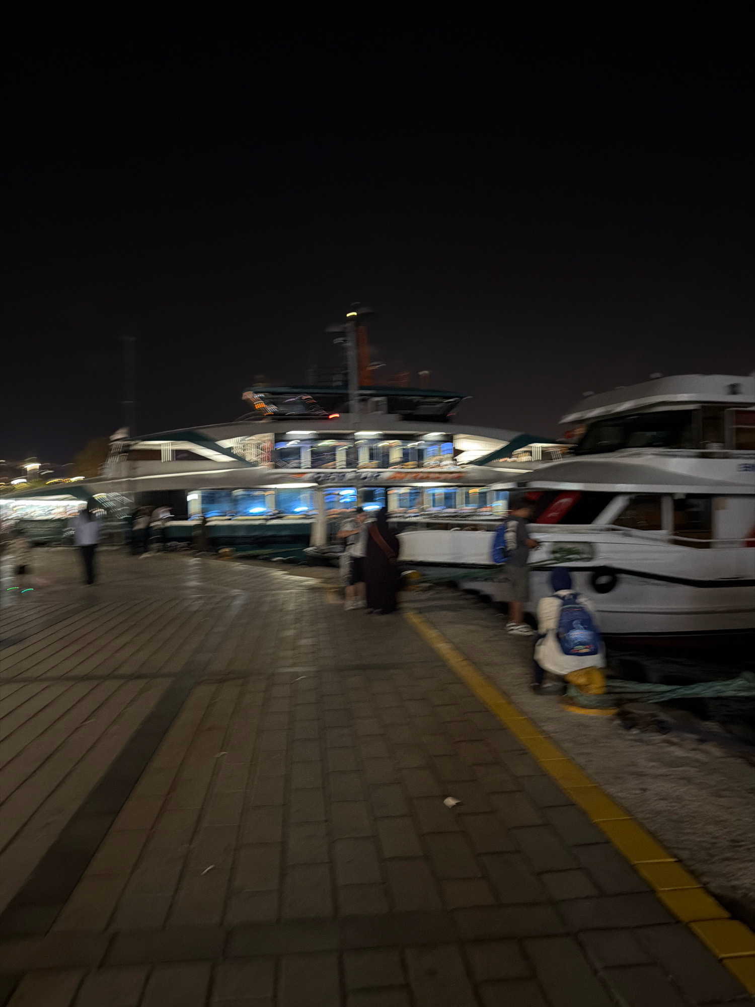 This is a nighttime photograph of a waterfront promenade in Üsküdar, Turkey, with several boats docked alongside. The promenade is paved with interlocking rectangular blocks, and a yellow line marks the edge closest to the water.
In the foreground, a person wearing a white top and yellow pants is sitting on the ground, facing away from the camera, with a blue backpack on their back. To their left, a person in a gray shirt and dark pants is standing, also with a blue backpack. Further to the left, a person in a white shirt and dark pants is walking away from the camera.
In the middle ground, two large passenger ferries are docked. The ferry on the left has multiple decks with illuminated windows, and appears to be green and white. The ferry on the right is predominantly white with dark windows, and has a Turkish flag visible on its side. Several blurred figures are visible on the decks of the ferries and on the quay between them.
The background is dark, indicating it is nighttime, with some distant lights visible. The overall impression is of a busy ferry terminal or docking area.