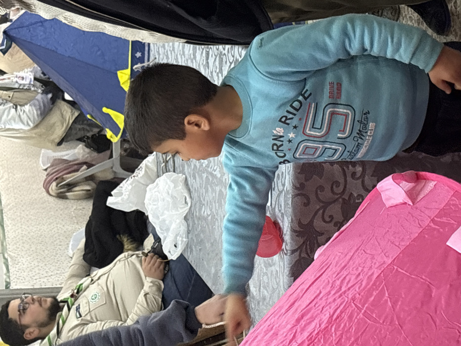 An indoor scene in Kafr Batna, Syria, depicts multiple individuals and objects within what appears to be a temporary shelter or community space. In the foreground, a young male child, with dark hair and wearing a light blue long-sleeved shirt bearing the text "BORN TO RIDE 95 Custom Motor Motorcycle Club," stands in profile facing left. His right arm is extended forward, pointing towards or touching a bright pink fabric play tent or structure, the base of which is visible in the lower left quadrant of the frame. A small red object, possibly a plastic cup, rests on a grey patterned tablecloth directly in front of the child's extended arm.
To the left and slightly behind the child, a male adult with glasses and a beard is seated. He is wearing a light tan uniform shirt with a green neckerchief and badges, indicative of a scout or volunteer organization. This individual is looking towards the right side of the frame, observing the scene. Behind him, a dark jacket is draped over a white plastic chair, and a white plastic bag rests on the table. Further back, another white plastic bag and a blue and yellow small tent are visible. A person wearing a light-colored jacket is partially visible behind the blue tent. On the far right, a segment of an adult's torso and lower body, clad in a dark jacket and olive-green pants, is visible, indicating another bystander standing near the child. The background consists of a light-colored, textured wall or partition.