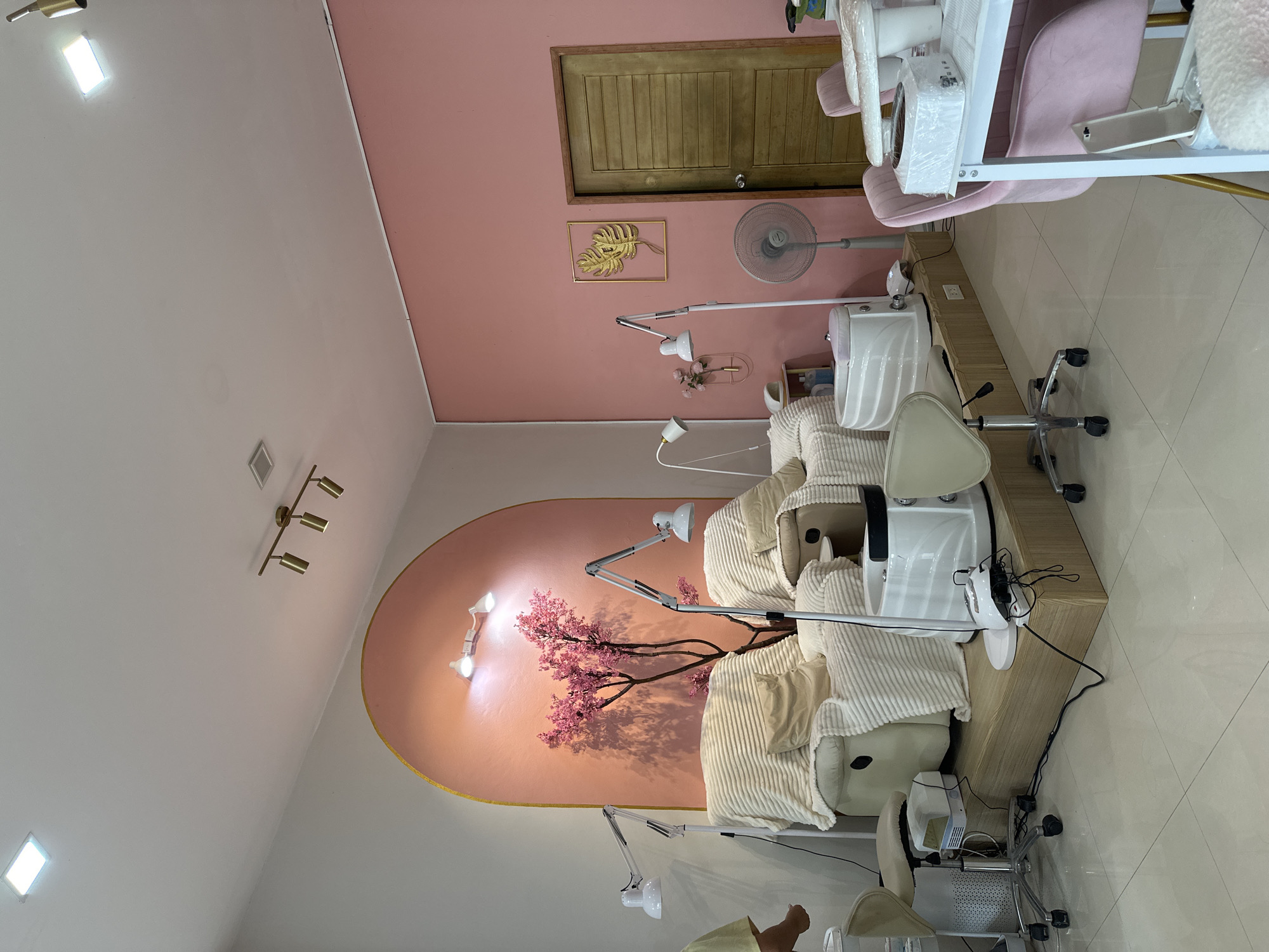 This is an indoor scene of a nail salon in Koh Phangan, Thailand. The room is decorated in shades of pink and white, with light-colored tile flooring. On the left, there are two cream-colored pedicure chairs, each covered with white blankets and cushions. In front of the chairs are white foot spa basins and adjustable task lamps. A decorative pink artificial tree is situated between the chairs, against a large arched wall section painted in a gradient of pink. In the background, a closed wooden door is visible on the right wall, which is painted pink. Above the door, a golden leaf-shaped wall decoration is mounted. A standing fan is positioned in front of the door. To the right of the fan, a white table with pink chairs surrounds it, set up for manicures. Several nail lamps and other manicure equipment are present on the table. The lighting is a mix of overhead ceiling lights and task lighting from the lamps.