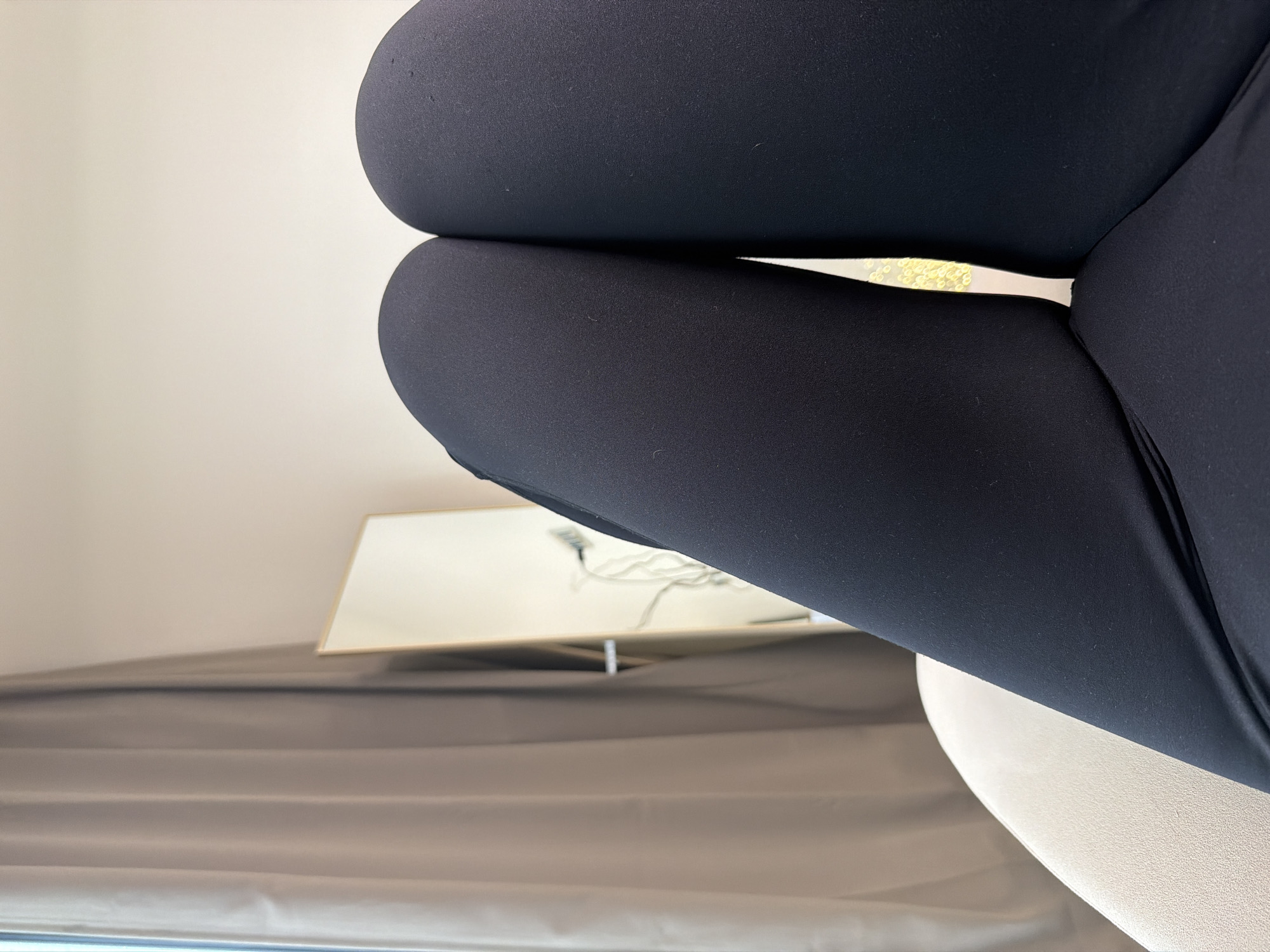A close-up shot focuses on a person's legs clad in black leggings. The legs are positioned above a partially visible white seat. In the background, there is a beige or light brown surface, possibly a bed or furniture. A square or rectangular white object with a gold rim rests on this surface, reflecting a distorted image of wires or cords. The overall setting appears to be indoors, possibly in a room with a simple and modern aesthetic.