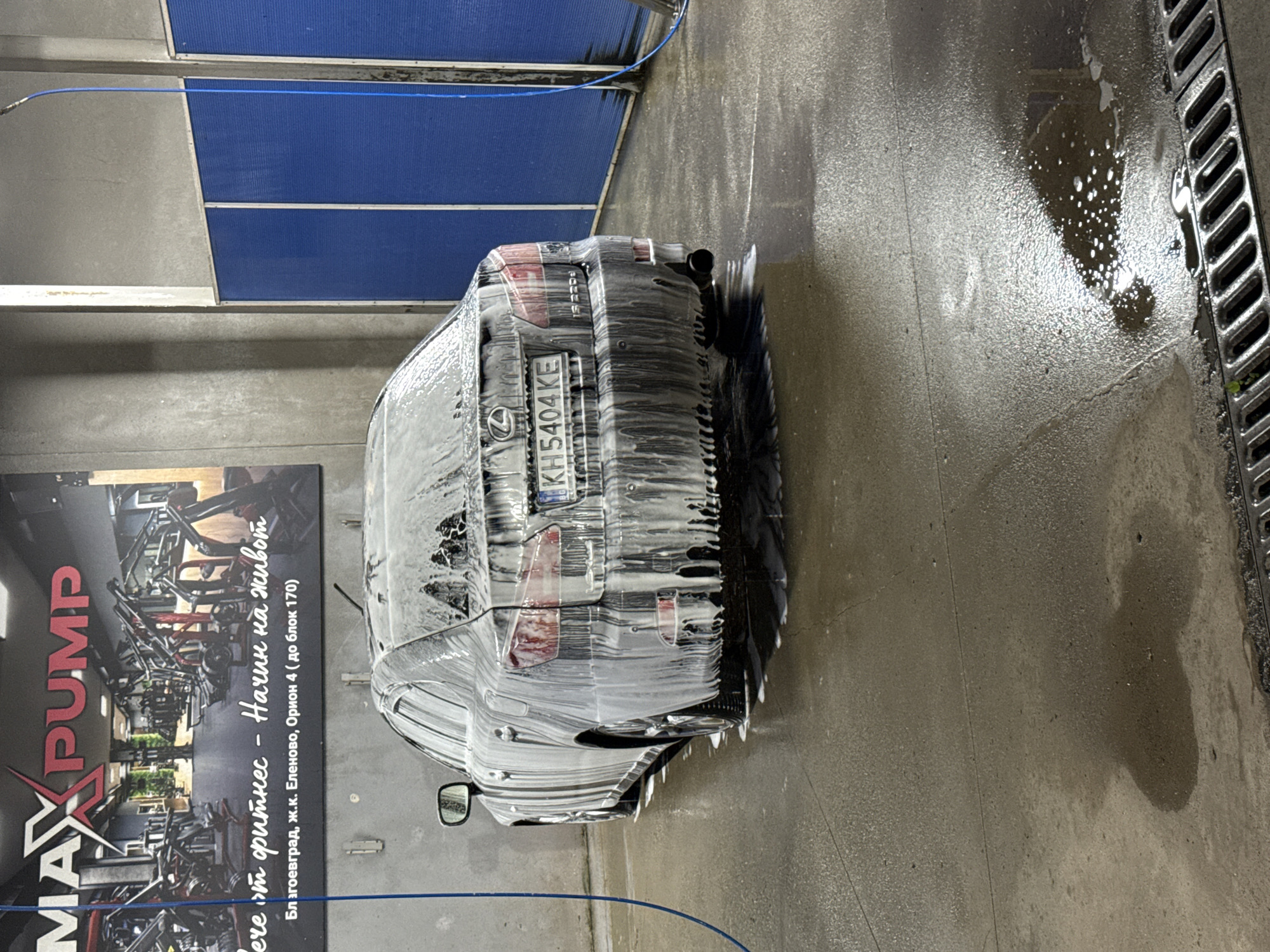 A dark-colored Lexus sedan is covered in white foam at a car wash. The car is facing away from the camera and is parked on a wet, concrete surface. The background features a large sign for "MAX PUMP" fitness center, with Cyrillic text below it indicating "Fitness - Start a new life" and the address "Blagoevgrad, J.K. Elenovo, Orion 4 (block 170)". Blue panels are visible on the right side of the frame, and a blue hose is hanging down from the top left. The floor is reflective due to the water and foam.