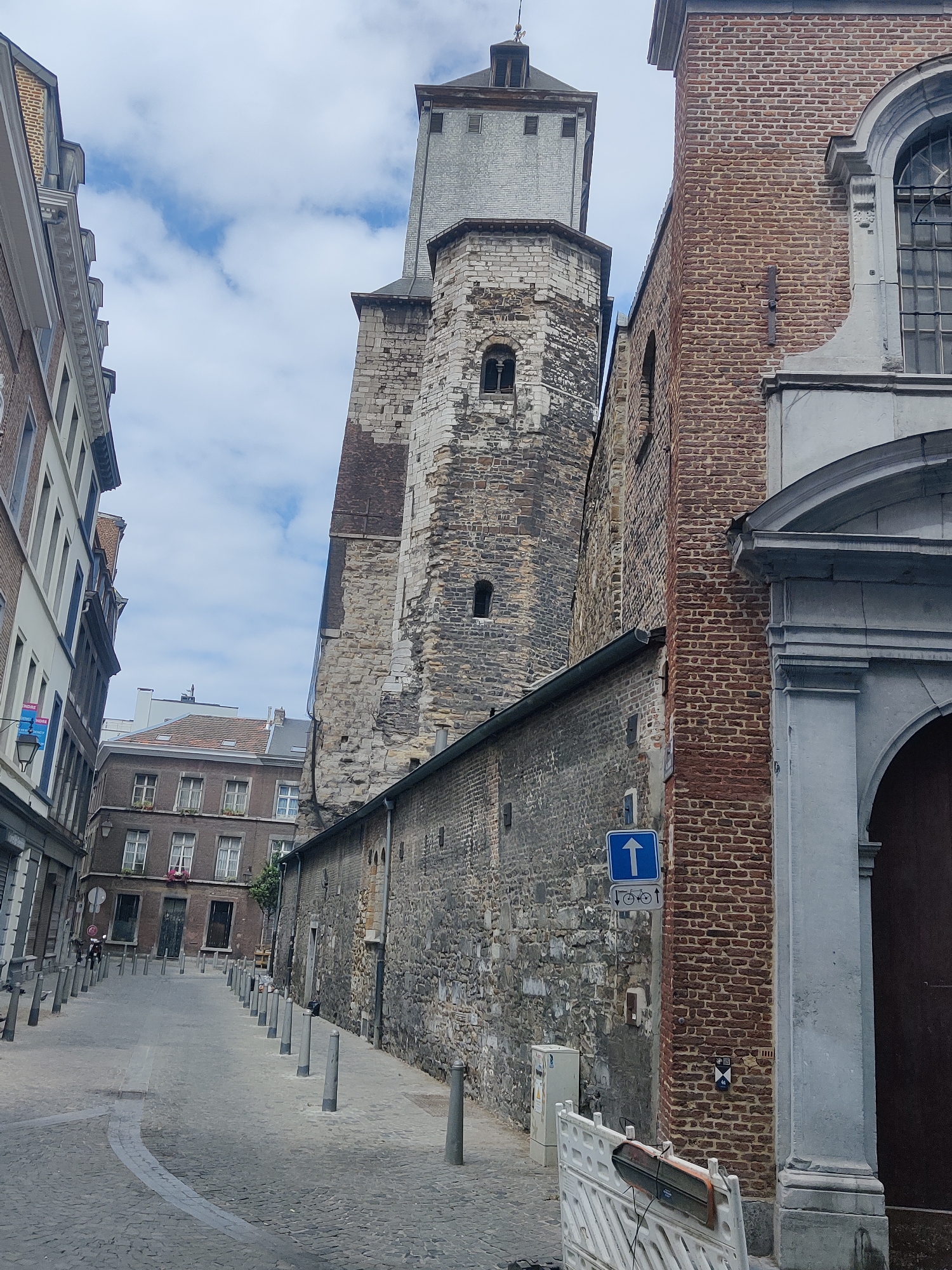 This image depicts a narrow street in the city of Liège, Belgium, with a tall stone tower dominating the background. The street is paved with cobblestones and lined with bollards. On the right, a building with a red brick facade features a large arched doorway. The street curves to the left, leading towards other buildings. The sky is partially cloudy.