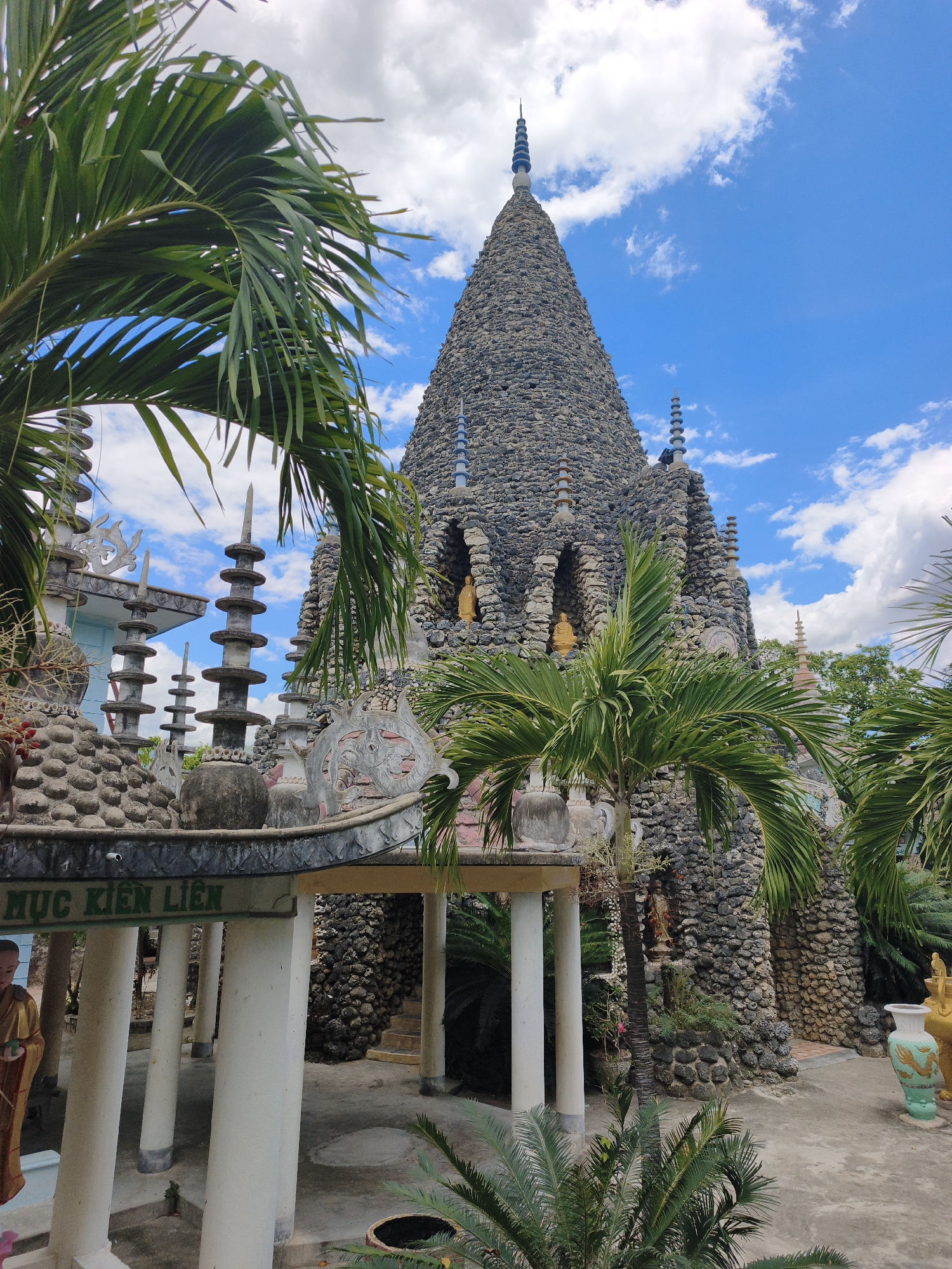 The image captures a view of a unique stone pagoda within the Tuan Thien Pagoda in Cam Ranh, Vietnam. The pagoda, constructed from numerous small, dark stones, rises to a pointed peak, adorned with tiered spires. Several smaller arched alcoves are visible in its structure, each housing a golden Buddha statue. The surrounding area is lush with tropical vegetation, including palm trees and cycads, their fronds framing the central structure. In the foreground, a covered walkway supported by white pillars is visible, with a sign bearing Vietnamese characters that read "DUC KIEN LIEN". The ground is paved with concrete. The sky is a vibrant blue with scattered white clouds. The overall scene depicts a serene and architecturally distinctive Buddhist temple complex set amidst a tropical environment.