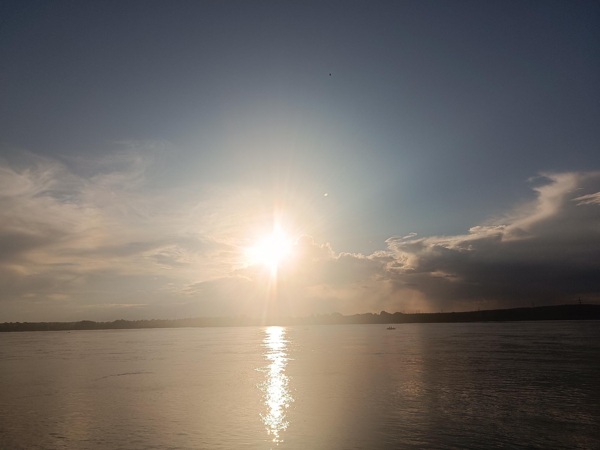 The image shows a wide river reflecting the bright sun, which is partially obscured by clouds. The sun's rays create a shimmering path across the water. The horizon is lined with a dark silhouette of trees and distant structures, suggesting a landmass. There are small boats visible on the water. The sky is a gradient of blue and grey, with wispy clouds on the left and darker, more substantial clouds on the right. The overall scene depicts a natural landscape at dusk or dawn, likely in the Chaikovsky area of Russia, given the presence of a wide river and the characteristic northern latitude sky.