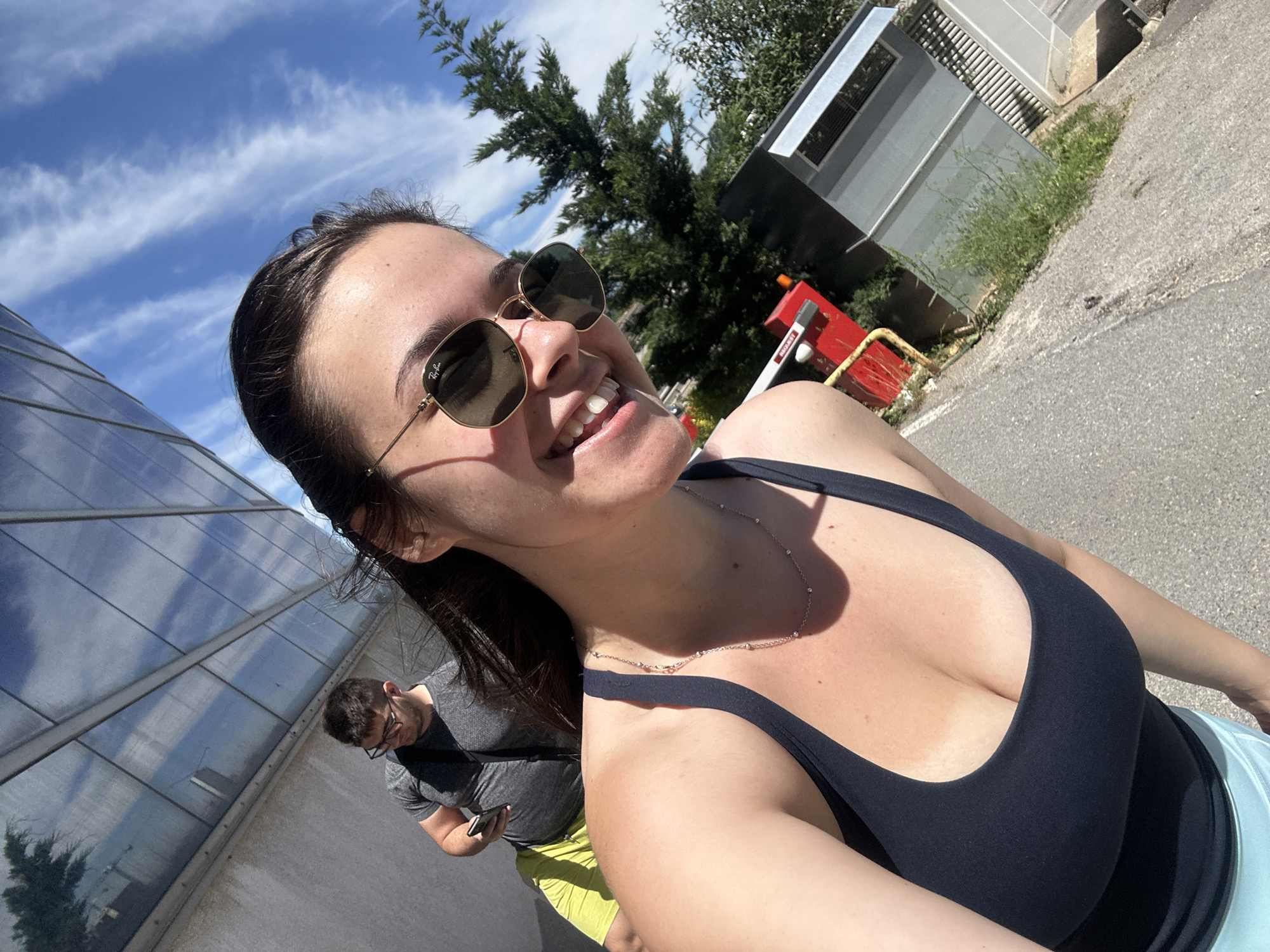 The image is a selfie taken from a low angle of a woman wearing sunglasses, a dark top, and a silver necklace. She is smiling. A man with glasses and a grey t-shirt is visible in the background, looking at his phone, partially obscured by a reflective glass building. The location appears to be outdoors on a paved area, with greenery and structures in the background. There is a building with a metal exterior and a red and yellow barrier nearby. Based on the surroundings and architectural details, this location is likely near the National Palace of Culture (NDK) in Sofia, Bulgaria. The day appears to be sunny with a blue sky and some clouds.