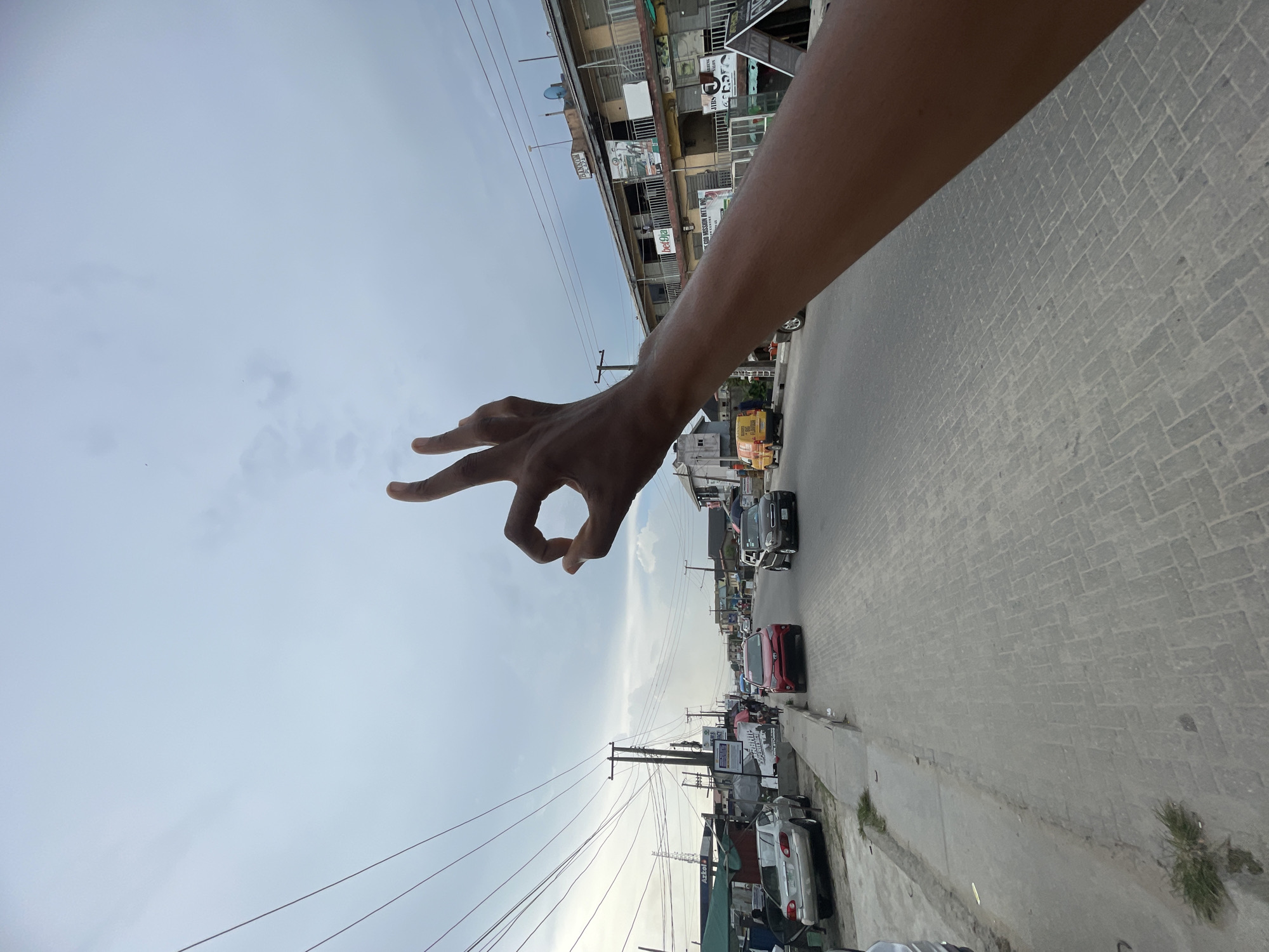 A human arm and hand, with dark skin, extends into the foreground from the upper right of the frame, making an "OK" gesture with the thumb and forefinger forming a circle and the other three fingers extended. The image depicts a street scene under a bright, partly cloudy sky during the daytime.
On the left side of the street, multi-story buildings line the road, displaying various commercial signs. Visible text includes "BANHAM HOTEL," "BEL9A," and partially "GOD MISSION INT. INC." Numerous utility poles and a dense network of overhead power lines are visible, extending along the street.
The paved road features a distinctive patterned surface, possibly brick or concrete pavers. Several vehicles, including cars and a yellow commercial van or bus, are present on the road, indicating traffic or parked cars. The scene suggests an urban or semi-urban environment, characteristic of a bustling street in Ado, Nigeria.