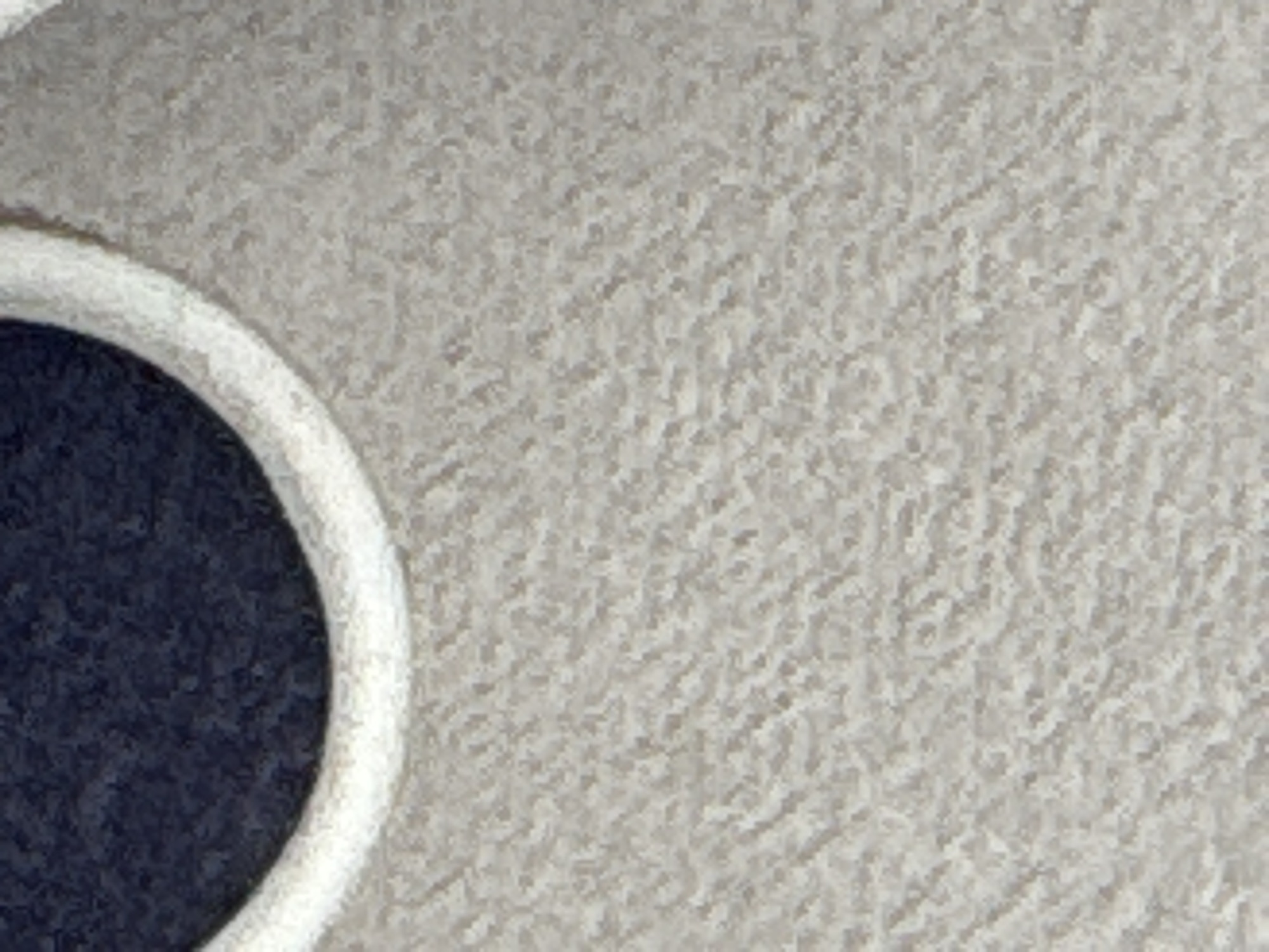 The image displays a close-up, abstract view of textured fabric. A dark blue circular area is bordered by a thick, off-white cord or trim. The surrounding fabric is a light, sandy beige with a noticeable, raised, woven texture that creates a speckled appearance. There are no discernible features to indicate a specific location in a city.