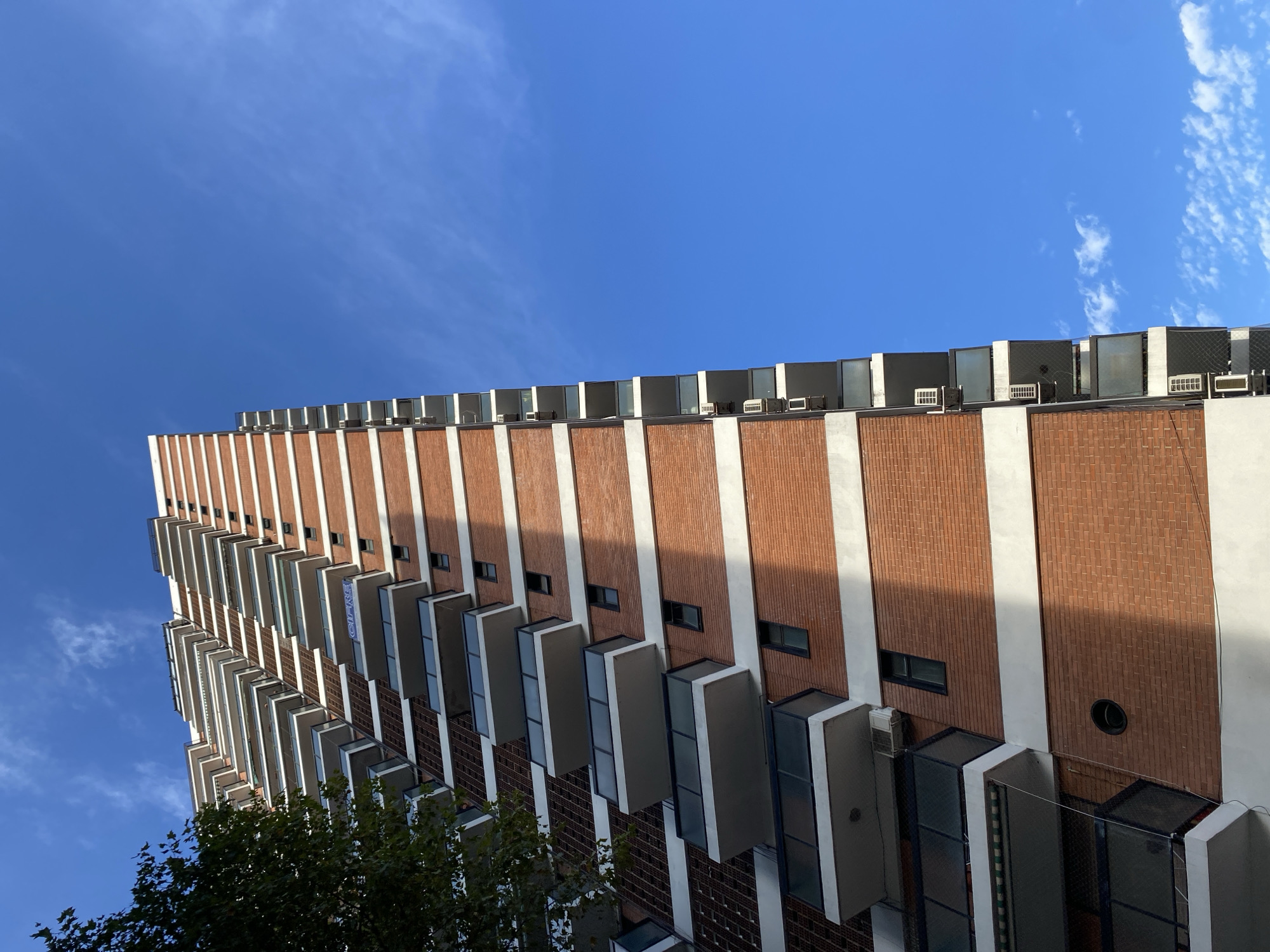 The photograph depicts a tall brick building with white vertical stripes in Buenos Aires, Argentina. The building features balconies with white frames and dark glass. On the roof, air conditioning units are visible. The sky is blue with some wispy clouds, and a tree with green leaves is visible in the lower left corner.