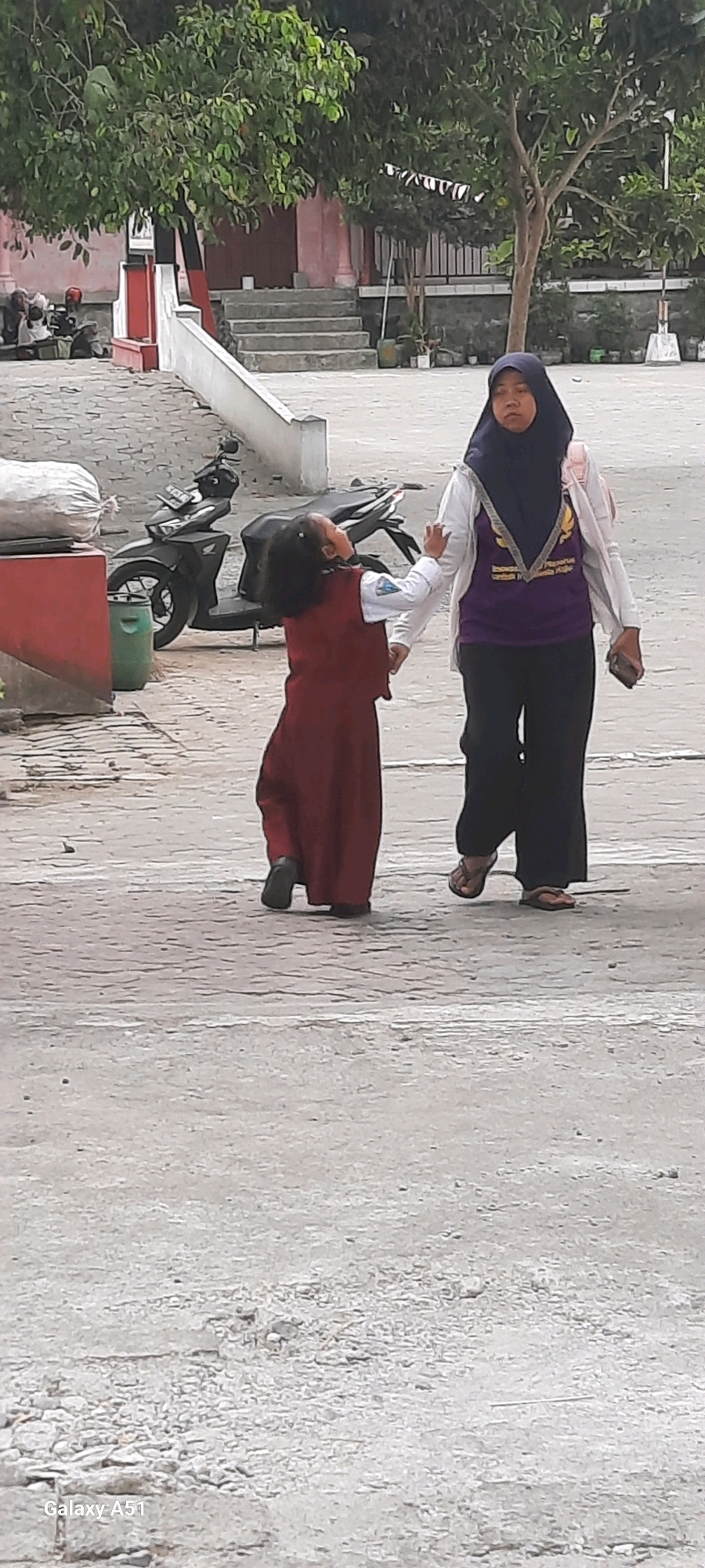 In the city of Tangkil, Indonesia, a woman wearing a navy hijab and a purple t-shirt with a white jacket walks alongside a young child. The child, dressed in a long maroon dress, reaches out towards the woman. They are on a paved outdoor area, possibly a schoolyard, with a motorcycle parked behind them to the left. In the background, there are stairs leading up to a building with trees and a fence. The ground is a textured gray pavement.