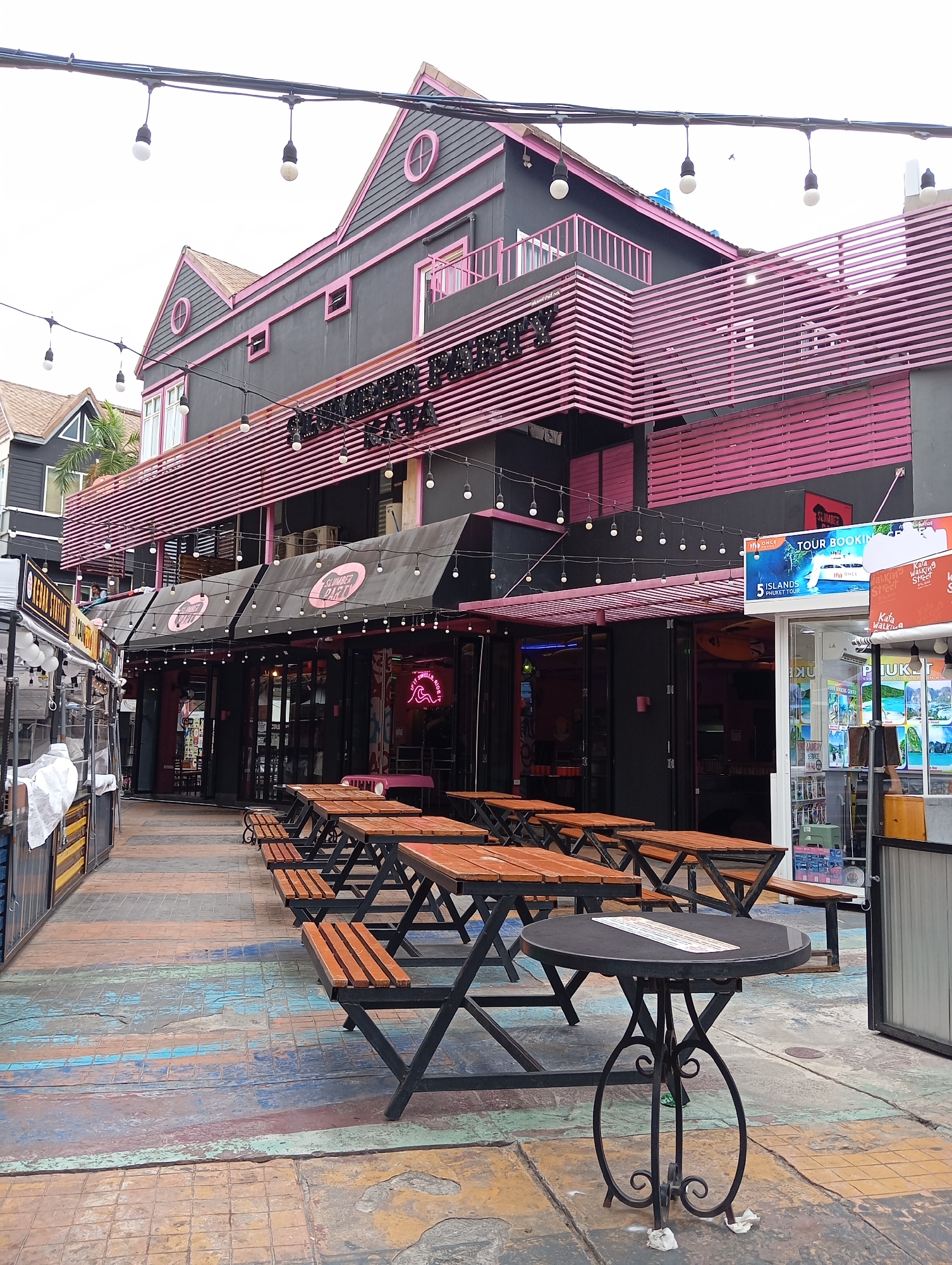 The image captures an outdoor seating area with multiple picnic tables and a round table in the foreground. The tables are made of wood and have black metal frames. The ground is paved with colorful, weathered wooden planks. In the background, a dark gray building with pink accents and decorative pink slats is visible. Above the building, strings of bare light bulbs hang. Signs for "Slumber Party" are visible above the entrance to the building, as well as a tour booking advertisement. The overall scene suggests a commercial area, possibly a street in Karon, Thailand, with shops and restaurants.