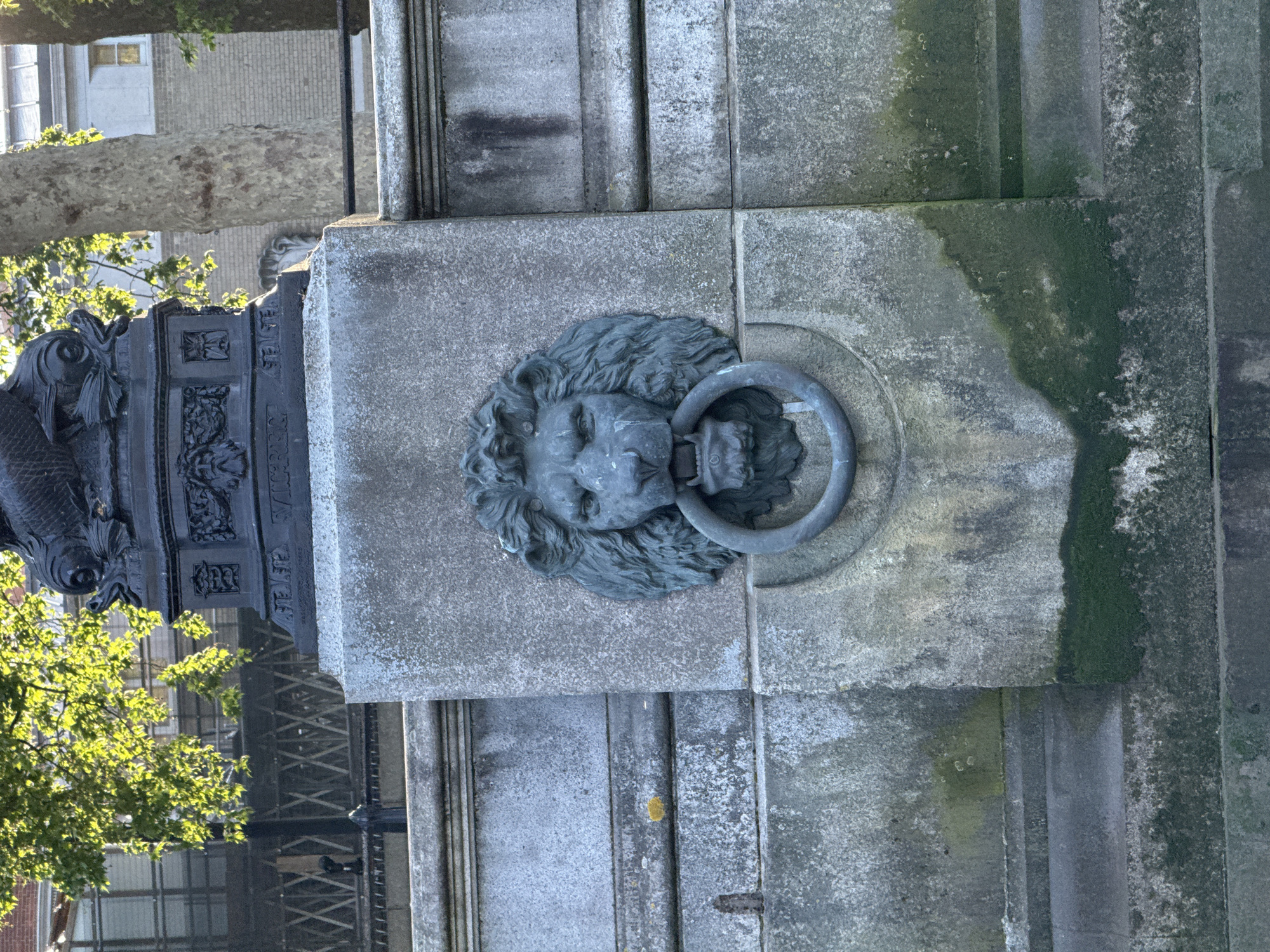 This is a close-up, horizontally oriented photograph of a stone monument in London, United Kingdom. The monument features a stone lion's head with a metal ring in its mouth, affixed to a weathered stone block. Patches of green moss grow on the stone. The base of a dark, decorative metal structure with fish motifs is visible to the left, partially obscured by greenery. In the background, glimpses of buildings and trees can be seen.