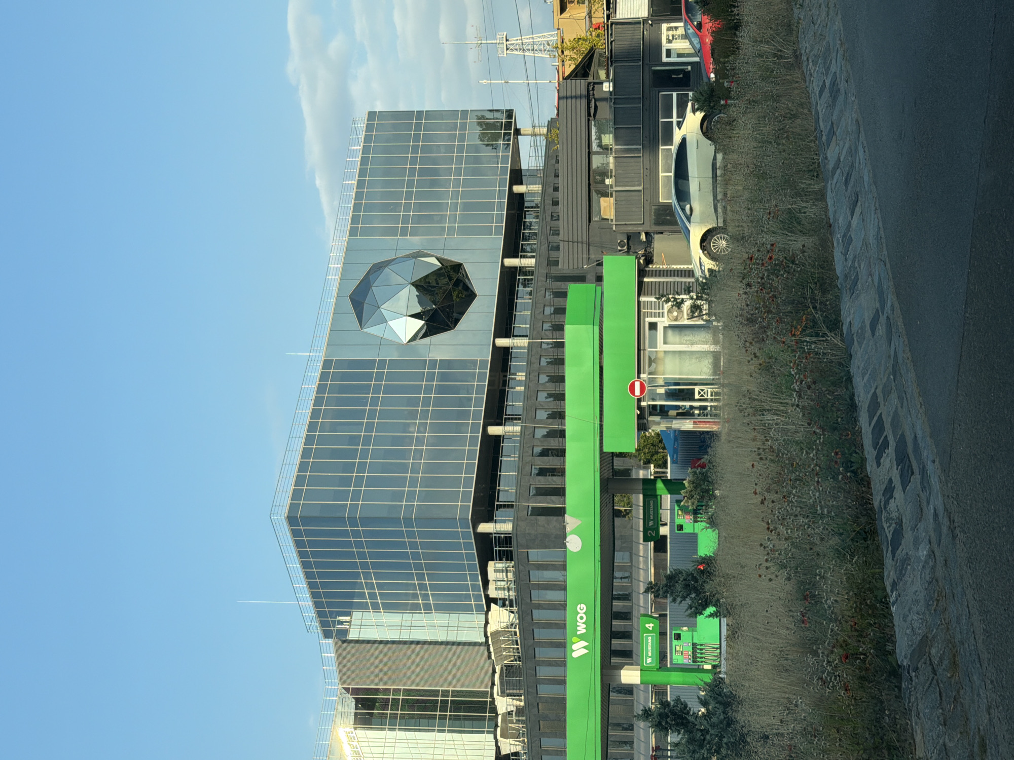 The image depicts a contemporary building complex with a prominent gas station in the foreground, located in the city of Kyiv, Ukraine. The building on the right side of the frame is a modern, multi-story structure with a facade largely composed of glass, featuring a distinctive, faceted geometric sculpture embedded within its upper section. To the left, a WOG gas station is clearly visible, identifiable by its bright green canopy and branding. Multiple fuel pumps are present, with signage indicating "MUSTANG" and numbers "4" and "2". Several trees and vegetation are scattered around the gas station and in front of the buildings. A light-colored car and a red car are parked near the gas station. The sky above is a clear blue with a few wispy clouds. The ground in the foreground consists of a paved road and a bordering area with cobblestone-like stones, transitioning into overgrown grass and wildflowers. In the background, a tall, lattice-like tower structure is visible.
