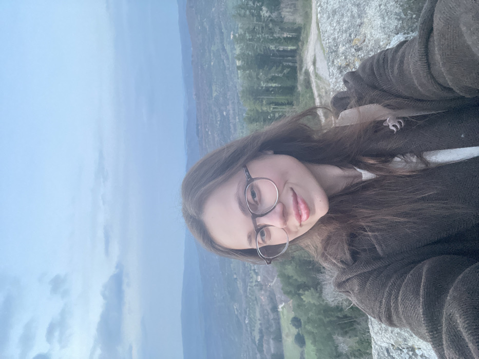 An image depicts a young adult female positioned in the foreground, facing the camera with a slight smile. She has long, dark brown hair, wears round-framed glasses, and is dressed in a dark brown textured coat over a light-colored top. Her left arm is raised, suggesting she is holding the camera for a selfie. She is situated on a grey, uneven rock surface, indicating an elevated position. The background reveals a panoramic view of rolling hills covered in green vegetation, interspersed with distant settlements under an overcast sky with scattered grey and blue clouds. The environment corresponds to a rural or natural landscape located in the city of Castel del Piano, Italy.