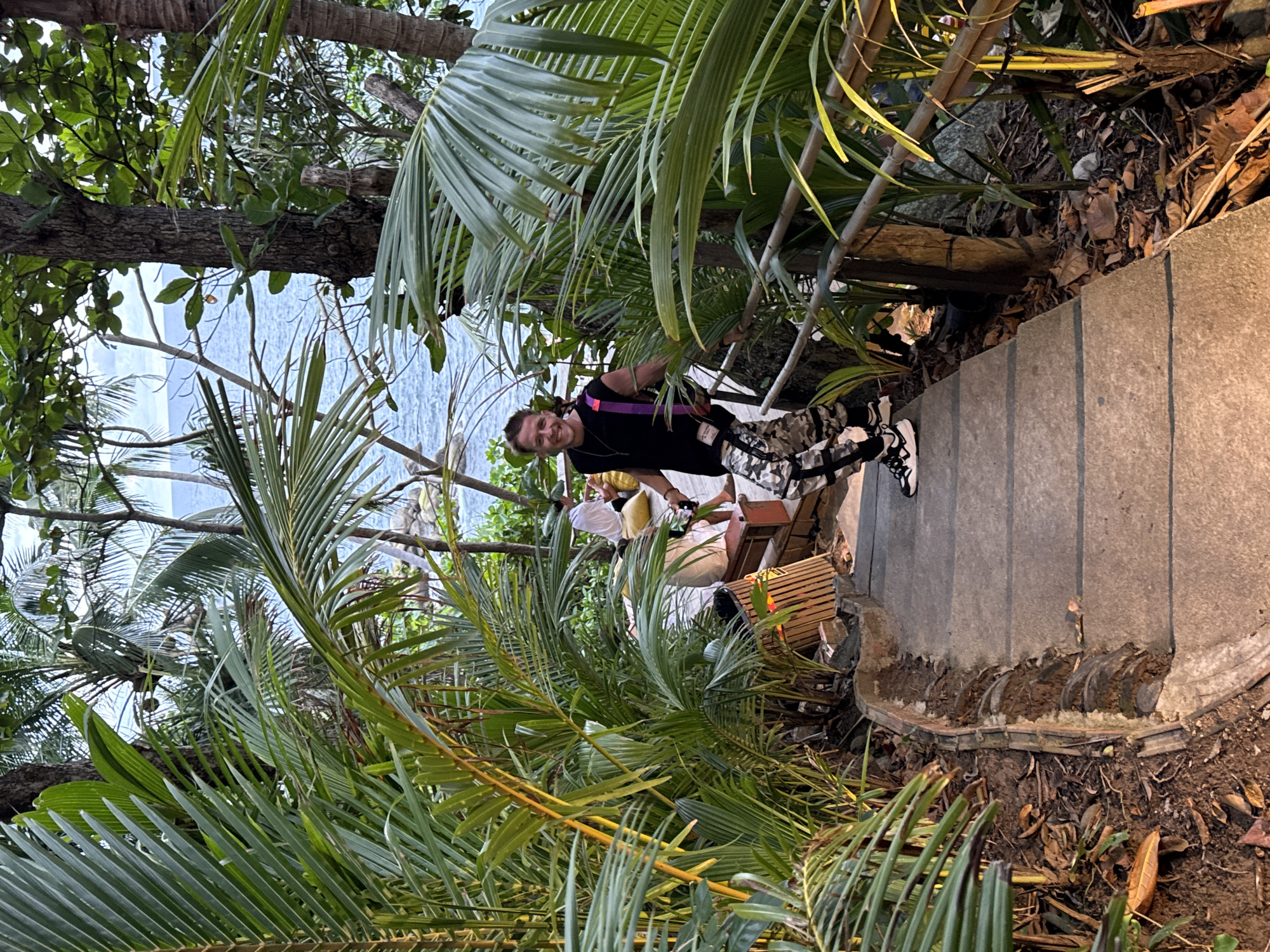 In Karon, Thailand, a young man with short blonde hair and a beard walks down a concrete staircase surrounded by lush tropical foliage. He is smiling and wearing a black sleeveless t-shirt, camouflage pants, and black and white sneakers. He carries a bottle in his left hand and a purple strap bag over his right shoulder. In the background, the ocean is visible between the palm trees, and a few people are seated on cushions in a dimly lit area. The staircase is lined with bamboo railings and vegetation.