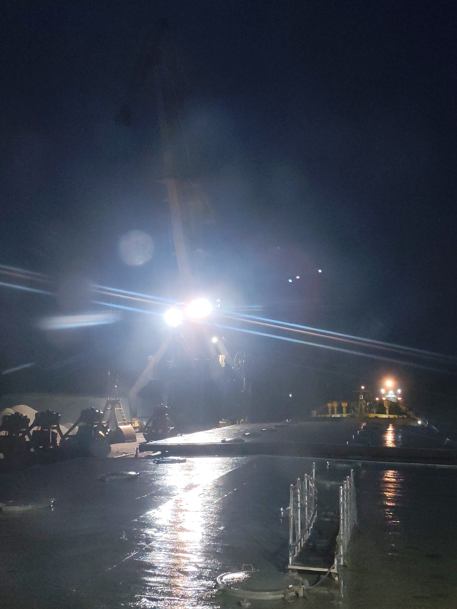 The image, taken at night, depicts a wet, reflective surface, likely a ship deck or industrial platform. A structure with bright lights dominates the center, possibly a crane or heavy machinery. The lights create significant glare and reflections on the wet surface. To the right, another structure is visible with illuminated lights. In the foreground, a metal railing is present. The overall scene suggests an industrial or maritime setting, possibly at night during or after rain. Location details are absent; the image alone does not confirm its location in Shalsky, Russia.