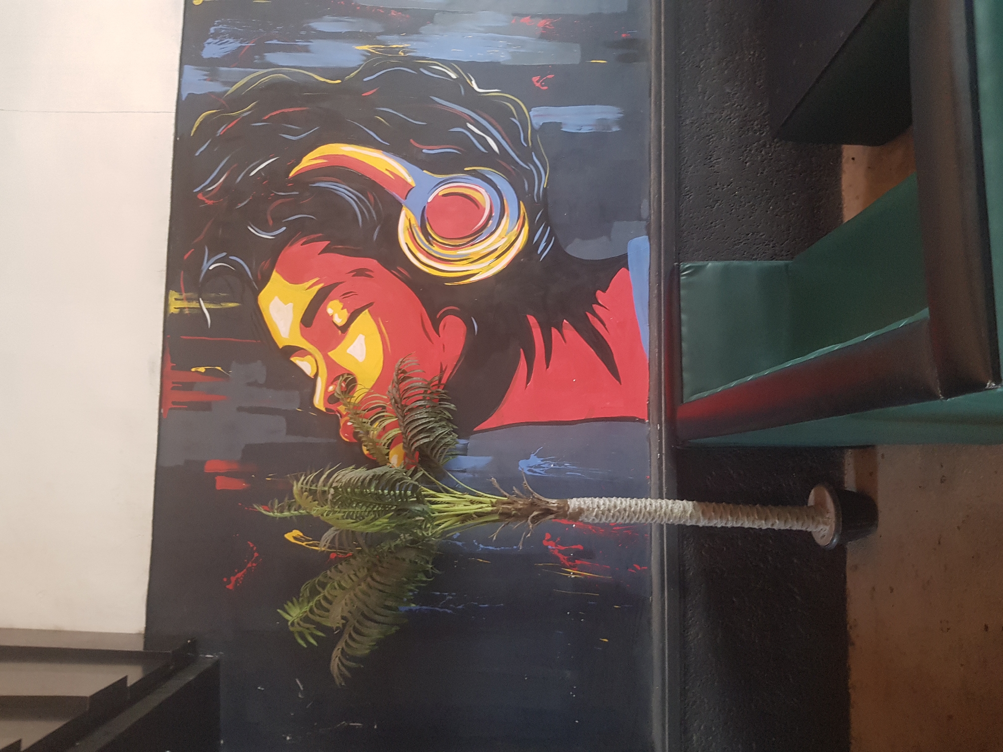 A large, vibrantly colored mural of a person wearing headphones is painted on a dark wall. The person's face is depicted in profile, with bold strokes of red, yellow, and blue. Splatters of red and yellow paint are visible on the wall around the mural. To the left of the mural, a small, artificial palm tree in a black pot stands on a rough, textured floor. In the foreground, a green and black upholstered bench seat is visible. The overall environment appears to be an indoor space, possibly a lounge or entertainment venue. The scene is located in Howayi, Nigeria.