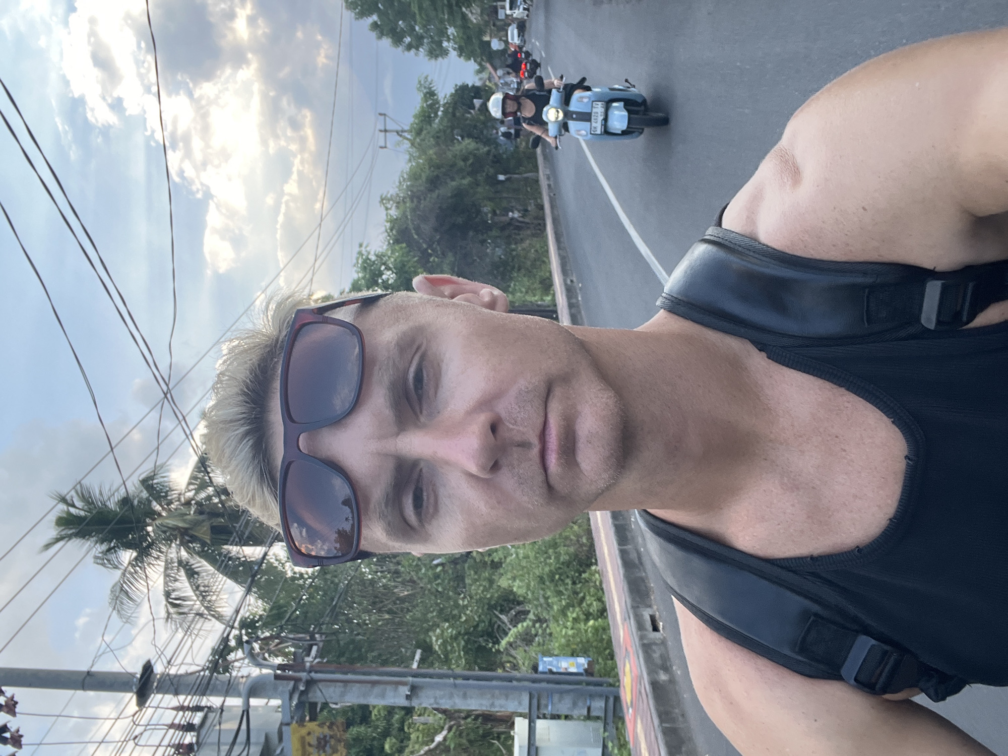 In a self-taken photograph, a man with blond hair and sunglasses rests on his head is the primary subject. He is wearing a black tank top and a black backpack. He appears to be riding a motorbike.
In the background, a woman wearing a helmet and a black top is riding a light blue scooter. She is on the same road. Several other motorbikes and cars are visible on the road further in the distance. The road is paved and has a white line marking.
The environment consists of lush green vegetation and trees on both sides of the road. Tall utility poles with many wires are visible on the left side of the image. Palm trees are also present. The sky is cloudy with patches of sunlight. The scene appears to be in Abang, Indonesia.