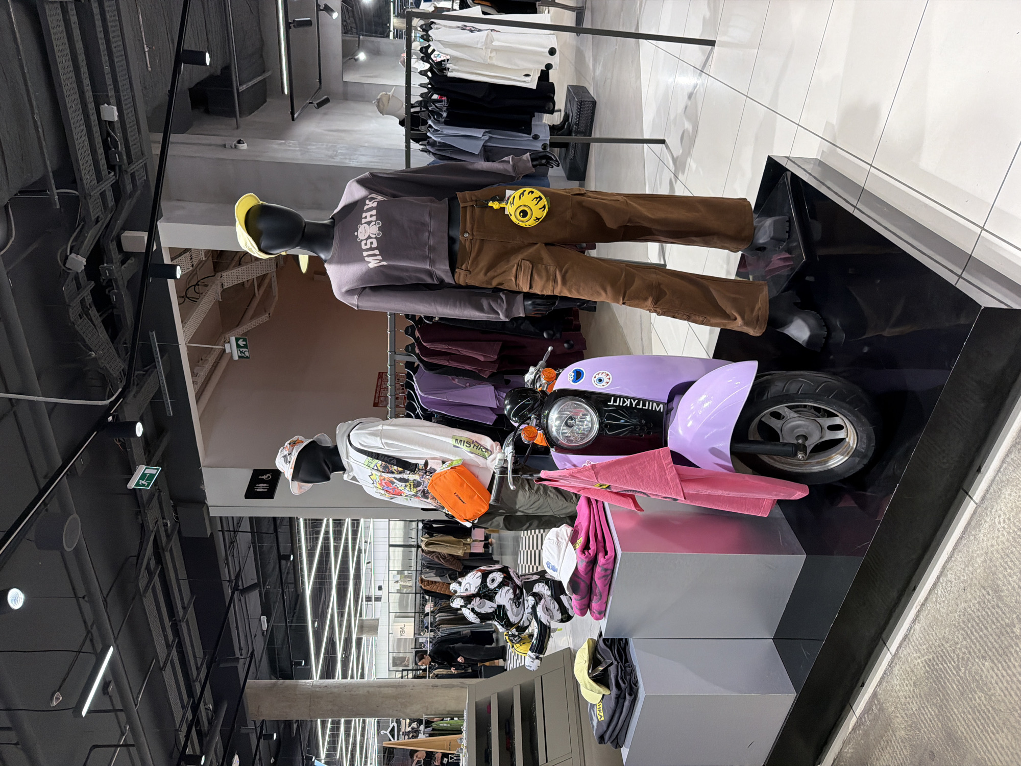 The image shows a retail store interior, likely in Khimki, Russia, based on the "WILDKRAFT" text on the purple scooter (a brand found in Russia). Two mannequins are dressed in clothing items. The upper mannequin wears a "MISHKA" branded sweatshirt, brown pants, and a yellow hat. The lower mannequin is dressed in a white hoodie, green pants, and carries an orange bag. A purple scooter is positioned between the mannequins. Clothing racks with various garments are visible in the background. The store has a modern design with exposed ductwork and lighting fixtures on the ceiling.