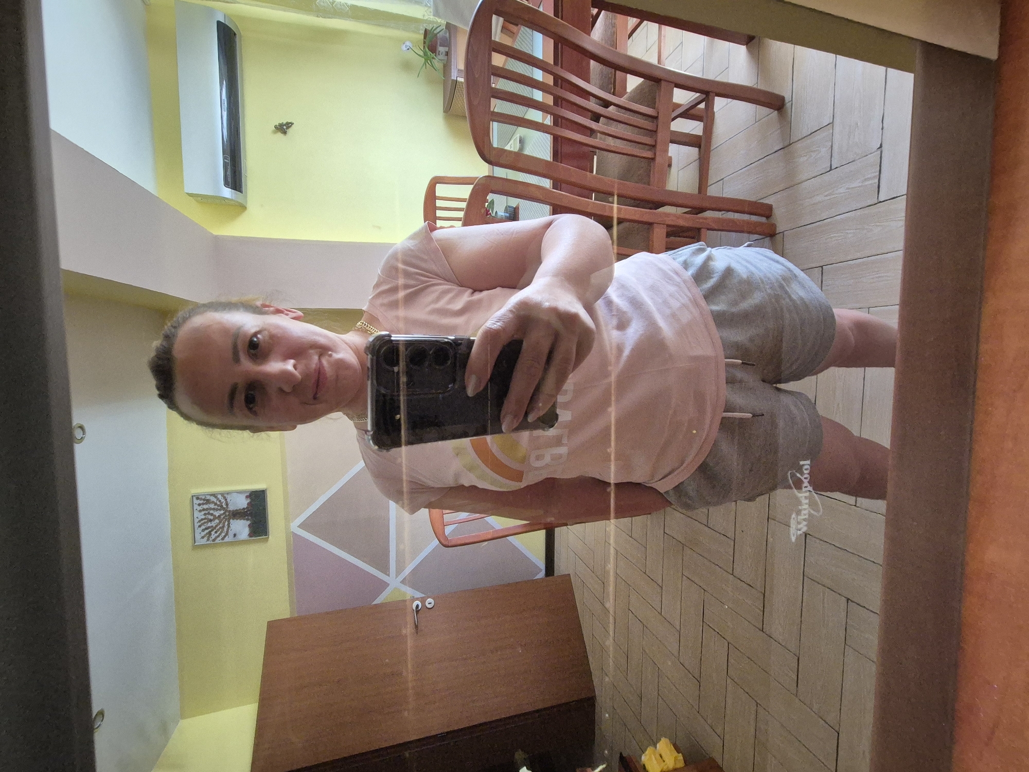 A mirror selfie taken by a woman in an indoor setting, potentially a home. The woman is fair-skinned with light hair, wearing a light-colored shirt and gray shorts. Her face is visible, and she is holding a smartphone in front of her to take the picture. The background includes a wooden chair, a wooden cabinet, and a wall with geometric patterns. The flooring appears to be tiled in a herringbone pattern. A framed picture hangs on the wall. There's an air conditioning unit mounted high on the wall. The image is somewhat distorted due to the mirror's angle. The location is Unknown, Unknown.