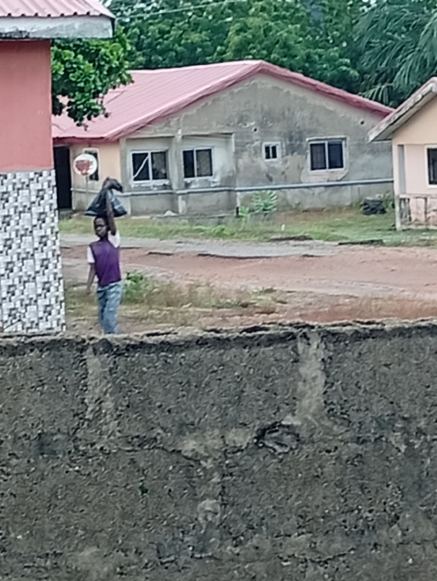 The image depicts a residential area in an unidentified city. A person is carrying a dark bag on their head while walking on a dirt path. Houses with red roofs are visible in the background. A concrete wall is in the foreground. The environment appears to be outdoors.