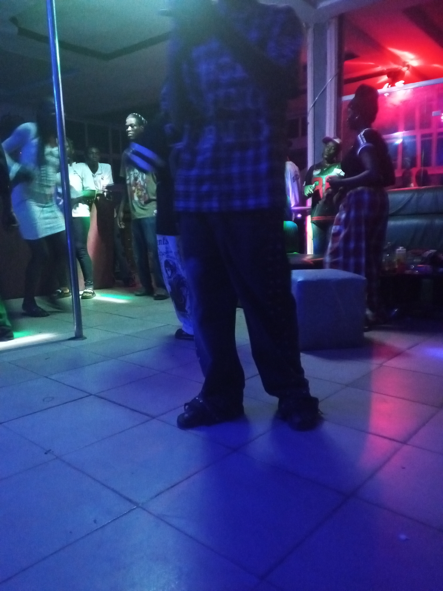 The scene appears to be inside a nightclub or bar, located in an unidentified city. The environment is dimly lit with blue and red colored lights. Several people are present, some standing near what appears to be a dance pole. A person in a plaid shirt and dark pants is prominently positioned in the foreground, obscuring some of the scene. In the background, people are seated on a couch and standing. The floor is tiled. The atmosphere suggests a social gathering or entertainment venue.