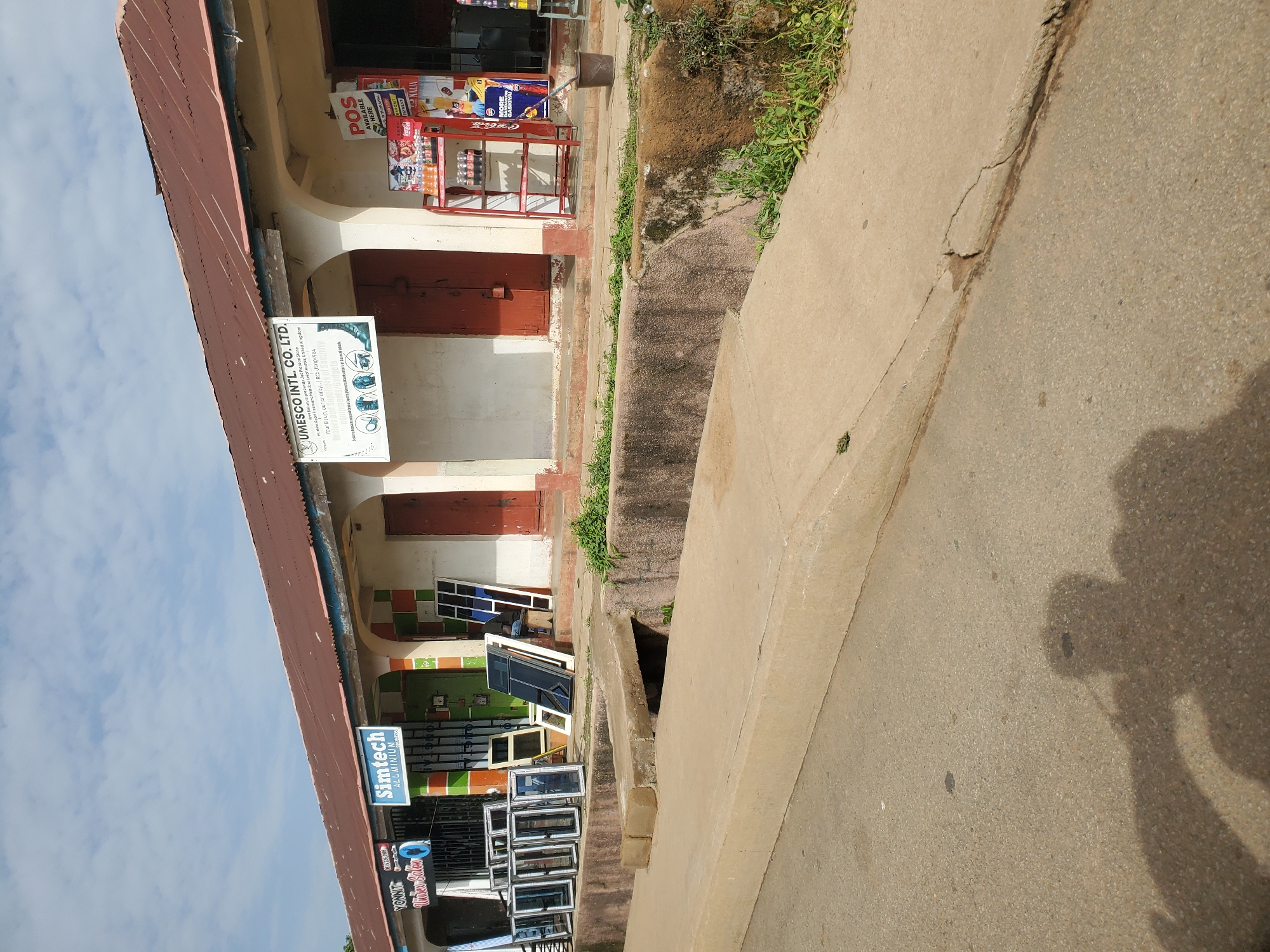 The image depicts a street scene in Bukuru, Nigeria, likely in front of a commercial establishment. The storefront features a corrugated metal roof, painted in a reddish-brown hue, with some rust and visible seams. The building has multiple arched doorways, some of which are closed with wooden doors painted red.
In front of the building, various aluminum window frames and glass panes are displayed, suggesting a business that sells or manufactures windows. One prominent sign reads "Simtech ALUMINIUM" with a logo. Another sign to the right advertises "UMESCO INTL. CO. LTD." and lists services like "Security Gadgets and Equipments/Hardware." Adjacent to this sign is a red metal display rack stocked with various beverages, including Coca-Cola, and other items. A dark bucket is placed on the ground near the display.
The foreground shows a concrete sidewalk and a road. A channel or gutter runs alongside the sidewalk, partially obscured by overgrowth. A shadow of what appears to be a person holding a camera is cast on the road, indicating the photographer's presence. The sky is partly cloudy.
