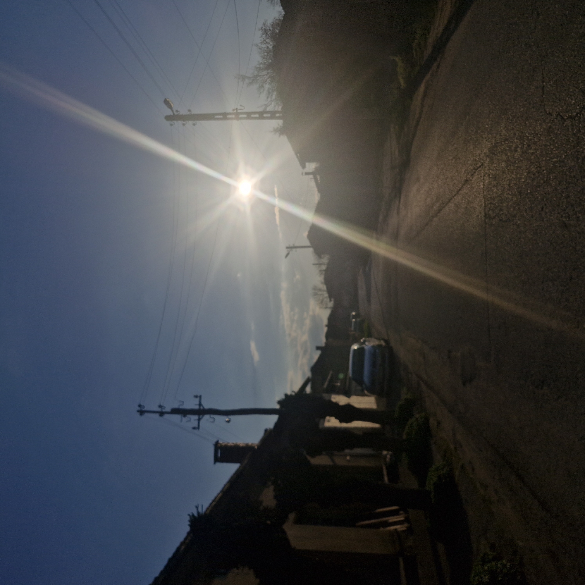 The image captures a street scene in Hirevo, Bulgaria, oriented vertically. The sun shines intensely in the upper-left quadrant, casting a bright flare across the sky and down the street. Overhead power lines run along the left side of the road, supported by wooden poles. A building with a chimney and visible roof is silhouetted in the foreground on the left. On the right side, the street surface shows texture and cracks, extending into shadows. Further down the street, two cars are parked. The foliage on the right and across the street appears dense.
