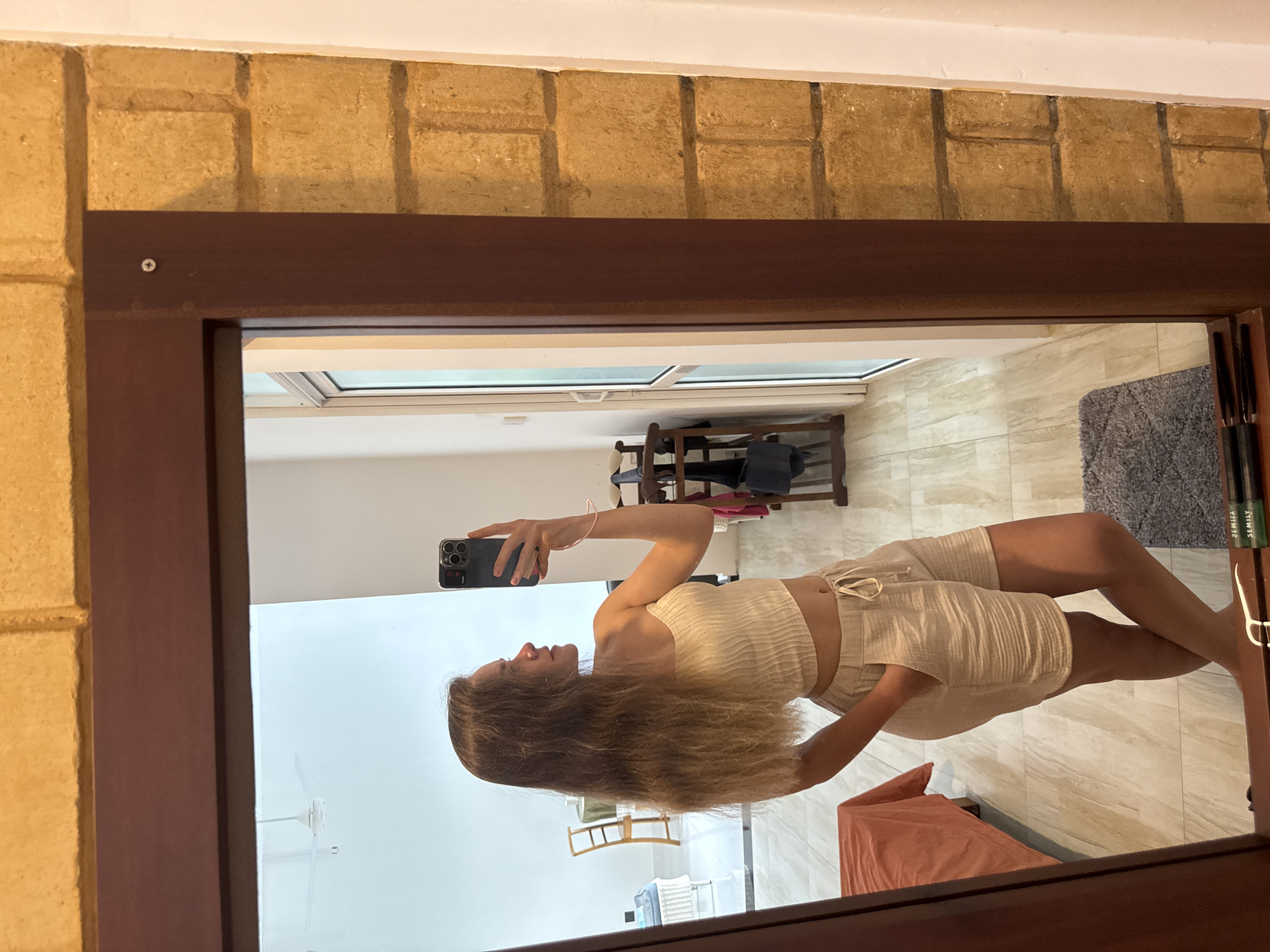 A woman is taking a selfie in front of a mirror in a room with tiled flooring, in Thalaramba, Sri Lanka. The mirror is framed with dark brown wood, mounted on a light brown brick wall. In the mirror’s reflection, the woman holds a smartphone and wears a cream-colored top and matching shorts. The room behind her includes a window, a wooden shoe rack, a gray rug, and a folded peach-colored item. The lighting is bright.