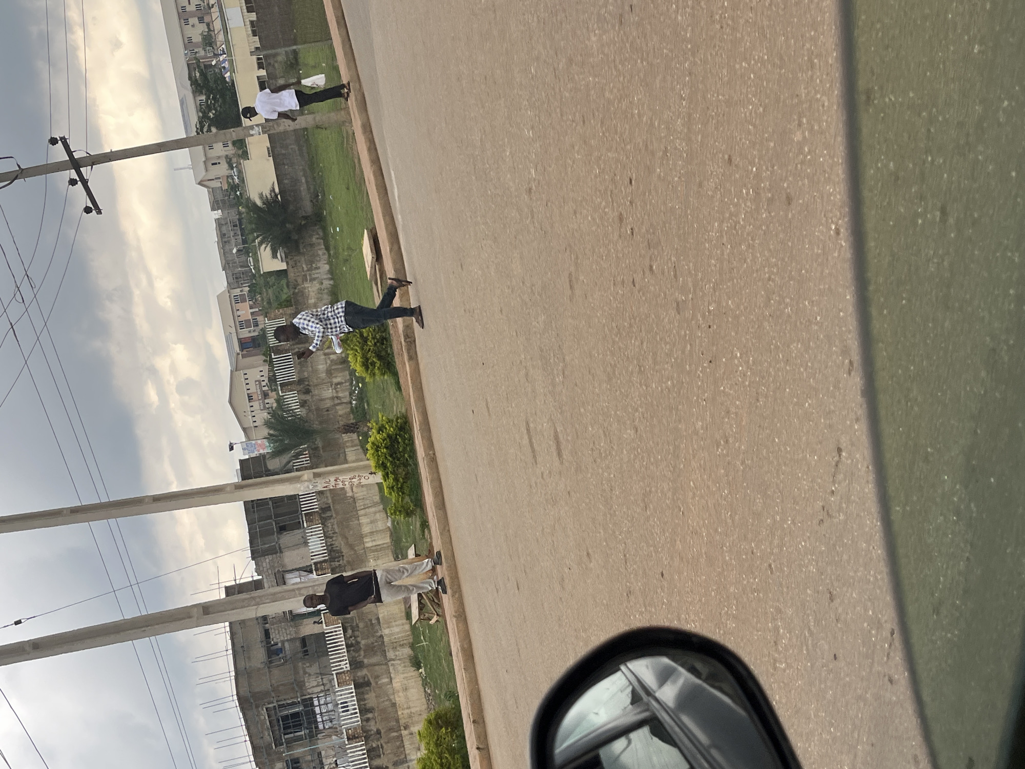 The image shows a street scene taken from within a vehicle, with a side mirror visible in the lower right. The road is asphalt with a curb separating it from a grassy area. Three people are walking along the side of the road. A person wearing a white top is in the background. Closer is a person wearing a checkered shirt. Farther in the foreground, a third person wears a dark top. Utility poles with wires are visible on the left. Buildings are in the background, with what appears to be construction occurring on at least one. The location is identified as Abuja, Nigeria.