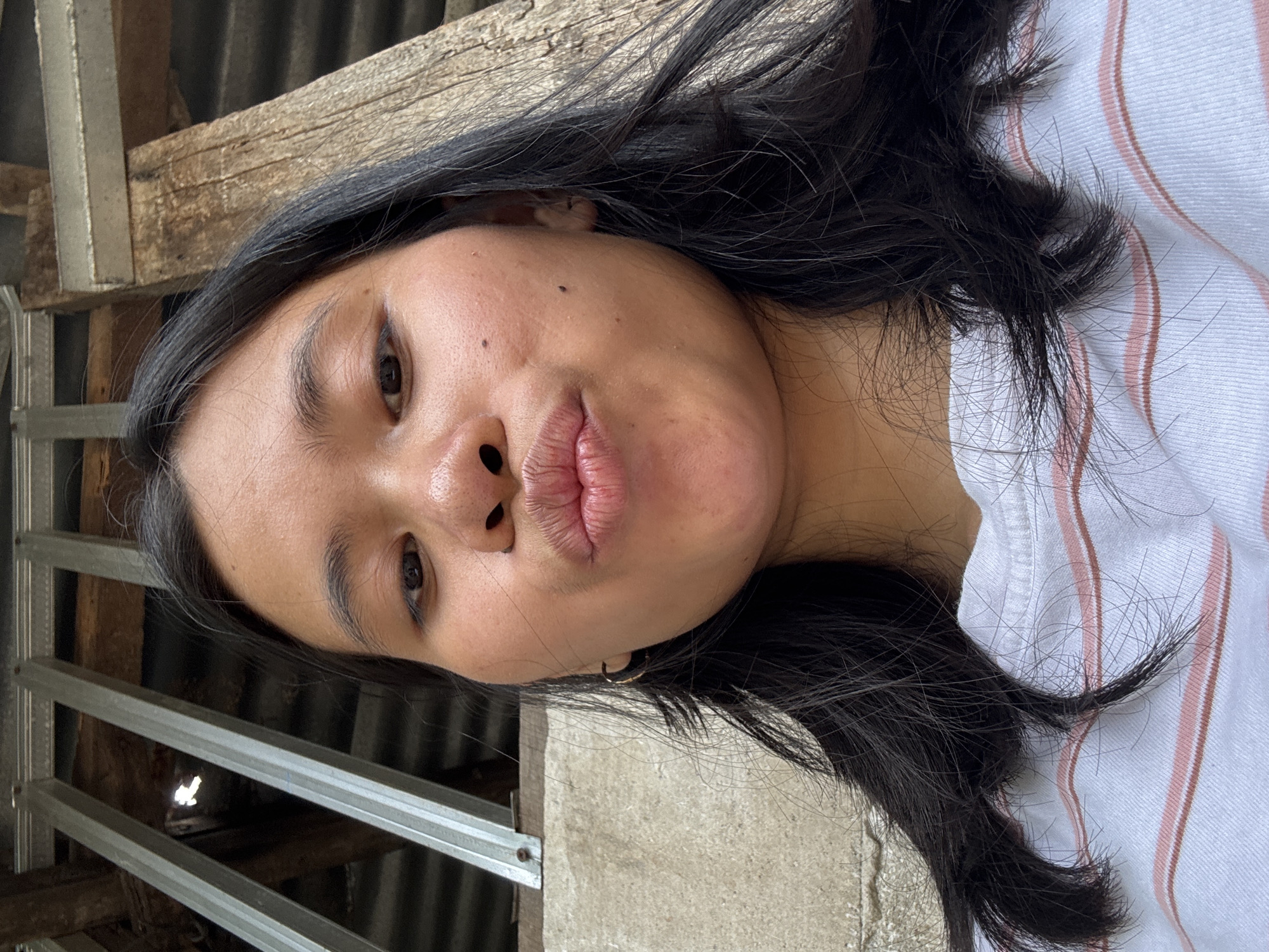 A low-angle, medium shot shows a young woman from the chest up, posing for a selfie outdoors. She has dark hair and is puckering her lips. She is wearing a white t-shirt with thin pink and white stripes. The background appears to be an outdoor setting with a rough concrete wall to the left and corrugated metal roofing with wooden supports above. The lighting is natural and appears to be daylight. The location is the city of East Jakarta, Indonesia.