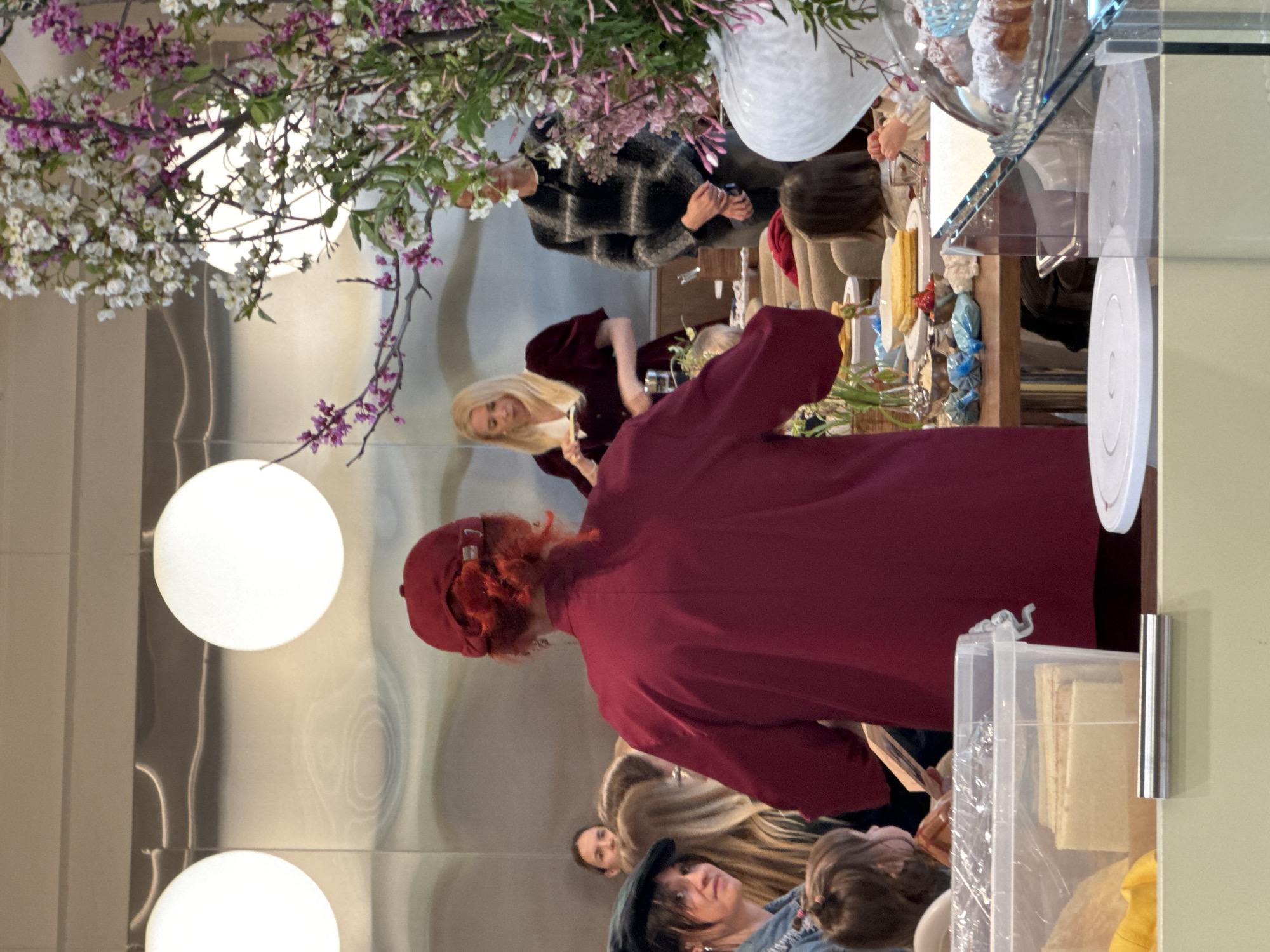 This indoor image captures a social gathering or event, likely in a cafe, shop, or exhibition space. The scene is illuminated by two large, circular white globe lights and features extensive floral decorations with white and pink/purple blossoms, possibly cherry or almond, creating a fresh, spring-like atmosphere.
In the foreground, a person with reddish-blonde hair, wearing a dark red long-sleeved top and a matching cap, stands with their back mostly towards the camera. Behind them, to the left, a blonde woman in a dark red velvet dress smiles engagingly, positioned behind what appears to be a serving or display counter. Further back, a person in a black and white plaid sweater holds a mobile phone. Several other individuals are visible throughout the space, some partially obscured, standing or seated, suggesting a diverse group of attendees.
The central feature is a counter or display table laden with items. A prominent glass tiered stand holds several pastries, possibly croissants, dusted with powdered sugar. Various other plates, bowls, and decorative elements are also present. On the right side of the counter, a clear plastic container holds light-colored contents.
The overall activity suggests an informal social event, perhaps a tasting, a product launch, or a casual gathering where food and drinks are served. The blonde woman's smile indicates a positive and welcoming mood. The image appears to be taken during the daytime. No specific text is visible in the scene. The location is contextualized as Khimki, Russia.
