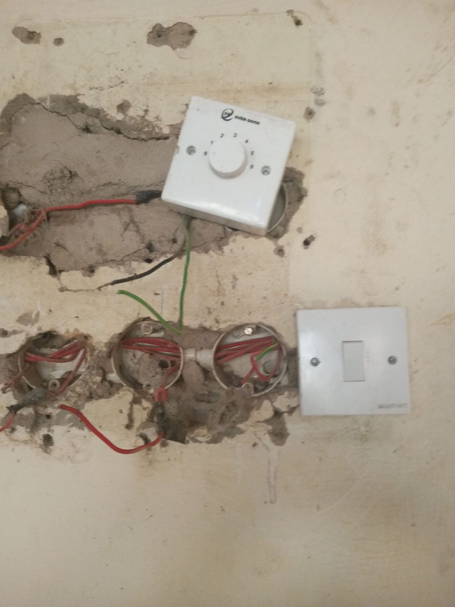 The image shows a damaged wall in Jalingo, Nigeria, with exposed electrical wiring. A white dimmer switch is mounted on the wall, and a white single light switch is also visible. Three circular electrical boxes, each containing red wires and some green wires, are embedded in the wall. The wall surface is cracked and rough, with evidence of plaster damage around the electrical installations. The overall scene suggests unfinished or damaged electrical work.