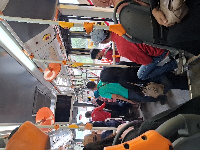 Inside a public transport bus, several passengers are visible. A man in a red shirt and jeans sits towards the front. Behind him, a person wears a green shirt. Overhead, the bus interior features numerous orange hand grips attached to the ceiling, along with white horizontal bars. Windows line the upper portion of the bus walls, letting in light. The scene depicts a typical commute with people seated and standing.