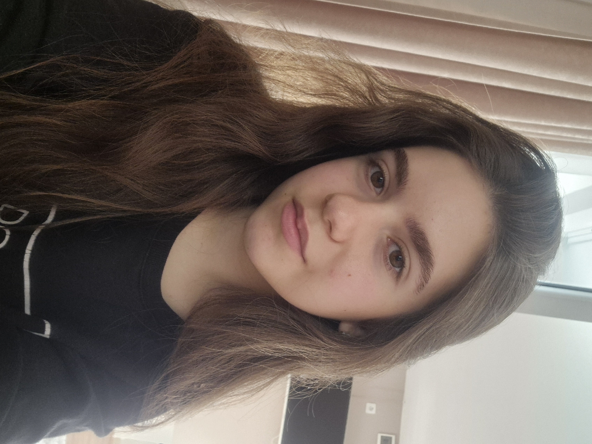 The image is a selfie of a young woman with long, wavy brown hair. She has light skin, brown eyes, and full lips. She is wearing a black shirt with white writing on the left side. The background includes a partially visible window with drawn blinds and a light-colored wall with a door and a framed picture. There is no indication of the location being Pavlikeni, Bulgaria.