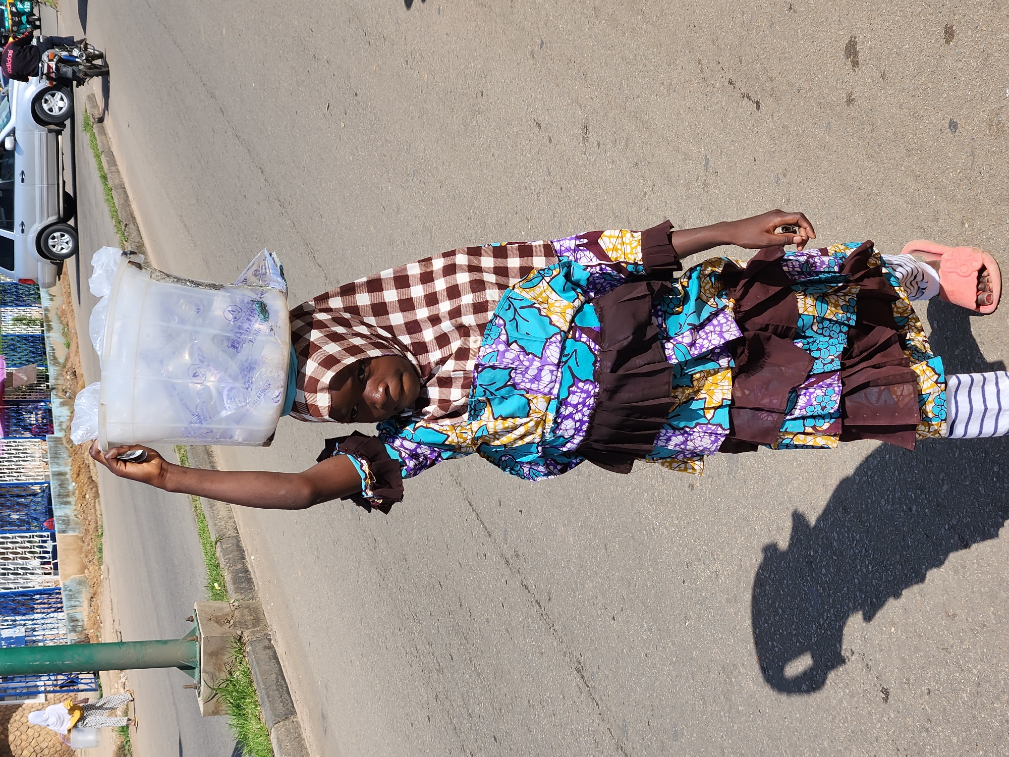 This image captures a street vendor in Jos, Nigeria, carrying a large container of water on her head. The vendor is a young woman dressed in a vibrant, patterned fabric and a brown and white checkered headscarf. She wears pink slippers on her feet, and her striped socks are visible. The water container appears to be made of clear plastic and is filled with bottled water.
The scene is set on a paved road, with a grassy verge and some blue and white fencing visible on the left side of the frame. In the background, a silver SUV and a motorcycle are parked, suggesting an urban or semi-urban environment. Another woman, also carrying water, is visible further down the road.
The lighting and the sharp shadow cast by the vendor indicate that it is likely daytime, possibly late morning or early afternoon, with bright sunlight. The image is taken from a slightly elevated angle, looking down on the vendor. The overall mood is one of everyday commerce and resilience, with the vendor focused on her task. There is no visible text in the image.