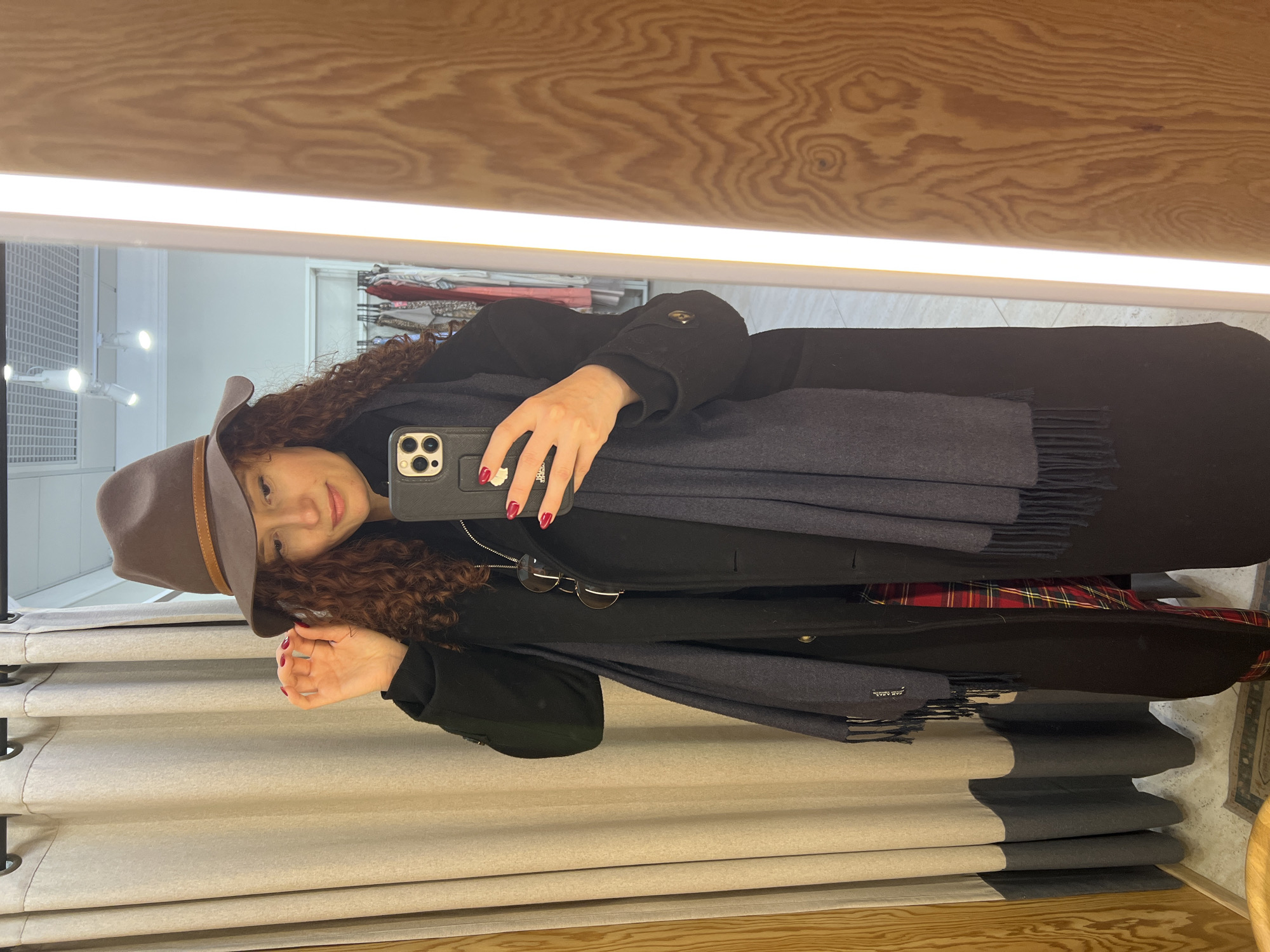 A woman with curly hair is taking a selfie in a mirror. She is wearing a brown fedora with a tan band, a black coat, and a dark blue scarf with tassels. She is holding a black smartphone with a camera in her right hand. She has red nail polish. Sunglasses are hanging from the front of her coat. The mirror is in a dressing room or retail setting with a light wooden frame at the top. White and gray vertically stacked curtains hang on the left. Garments are on a rack behind her to the left. It is not possible to determine the city of Saint Petersburg, Russia.