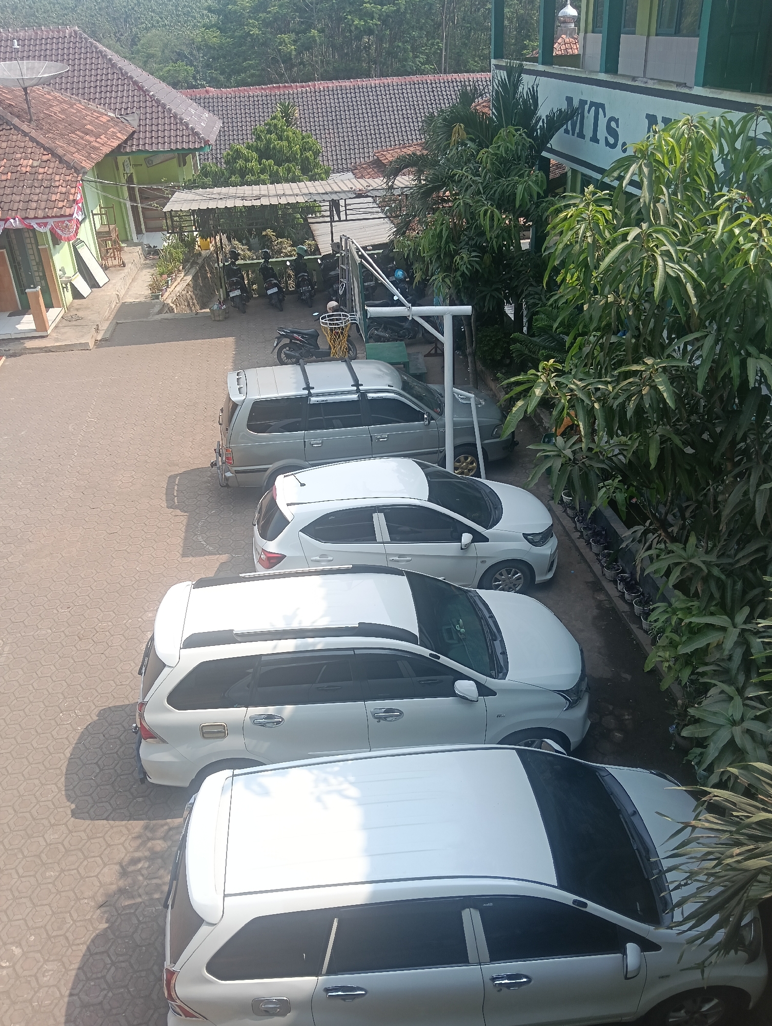 In the city of Adinuso, Indonesia, an outdoor scene captures a parking area and a building. A line of four white passenger vehicles is parked on a patterned concrete surface. Closest to the viewer is the rear of a white SUV, followed by another white SUV, and then two white sedans. To the left of the parked cars, a portion of a light green building is visible with a red-tiled roof. Further in the background, more red-tiled roofs and lush green foliage suggest a residential or educational setting. On the right side of the image, a larger building with a green facade and white trim features the text "MTS. N" in large white letters. A variety of trees, including those with broad leaves, flank this building and extend into the background. Several motorcycles are parked under a covered awning near the building. The sunlight casts shadows on the ground, indicating it is daytime.