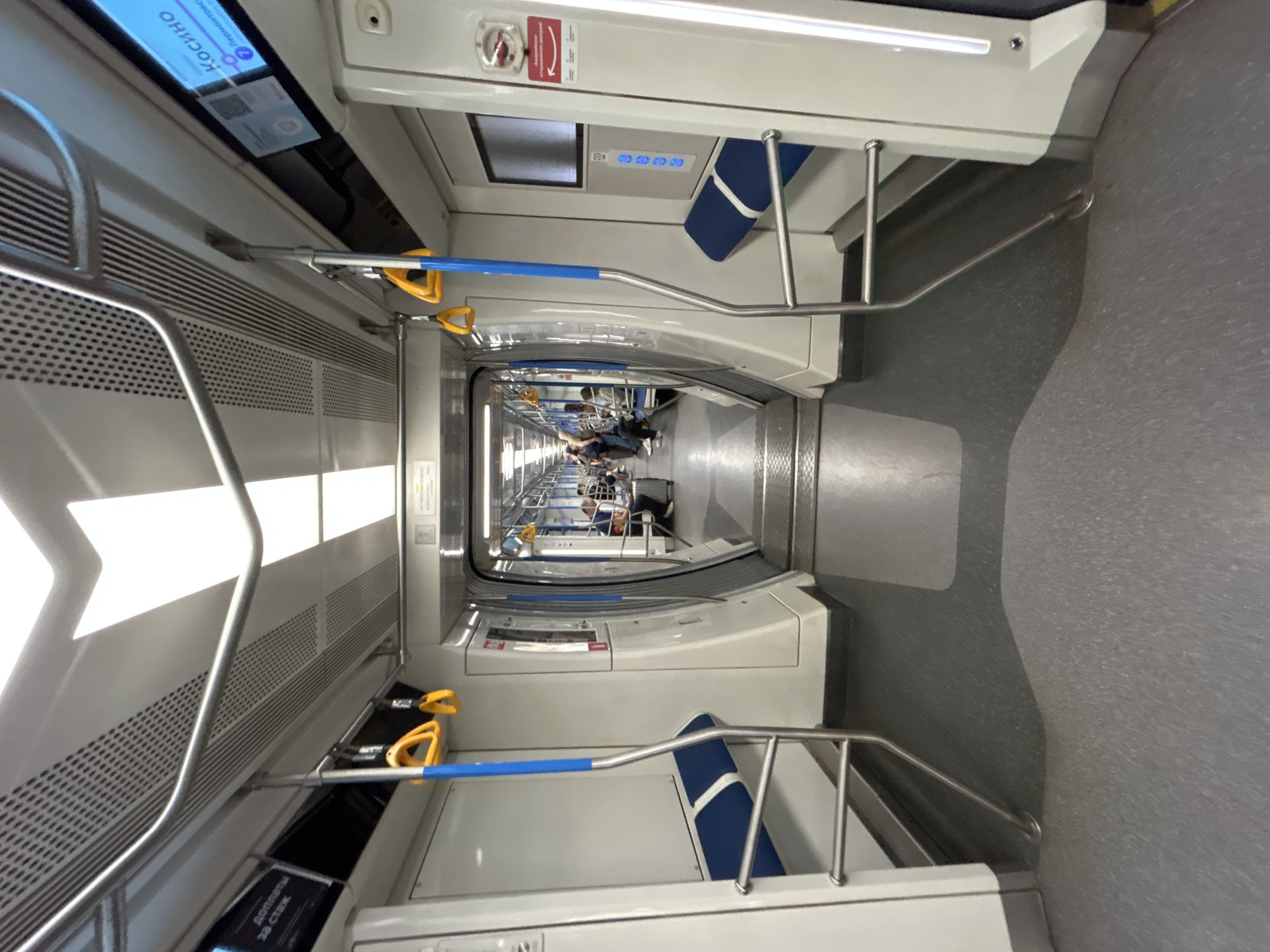 This is a low-angle, eye-level, wide-angle shot of the interior of a modern, clean subway car, possibly in Moscow based on the Cyrillic text visible on screens. The perspective is looking down the length of the car, with the seating areas and connecting passageway to other cars visible. The walls and ceiling are white and feature integrated lighting. There are blue and yellow handrails for passengers. The floor is a dark grey, textured material. Some passengers are visible seated and standing in the distance, with luggage near one passenger. The overall impression is one of a functional, well-maintained public transport vehicle.