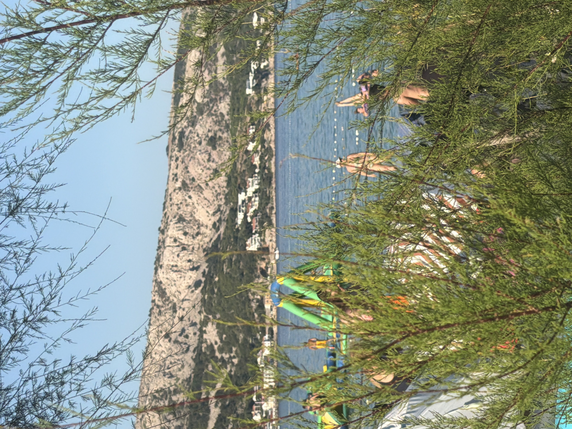 The image shows a sunny day at a beach in Općina Baška, Croatia. In the foreground, lush green branches of a tree obscure the view of the beach and the water. Beyond the foliage, the turquoise sea is visible, with several people enjoying various activities. To the left, an inflatable water park structure is partially submerged in the water, with a few individuals interacting with it. Further back, the shoreline is lined with buildings and houses, nestled against a backdrop of a rocky, green-covered hill. The sky is a clear blue, indicating fair weather.
