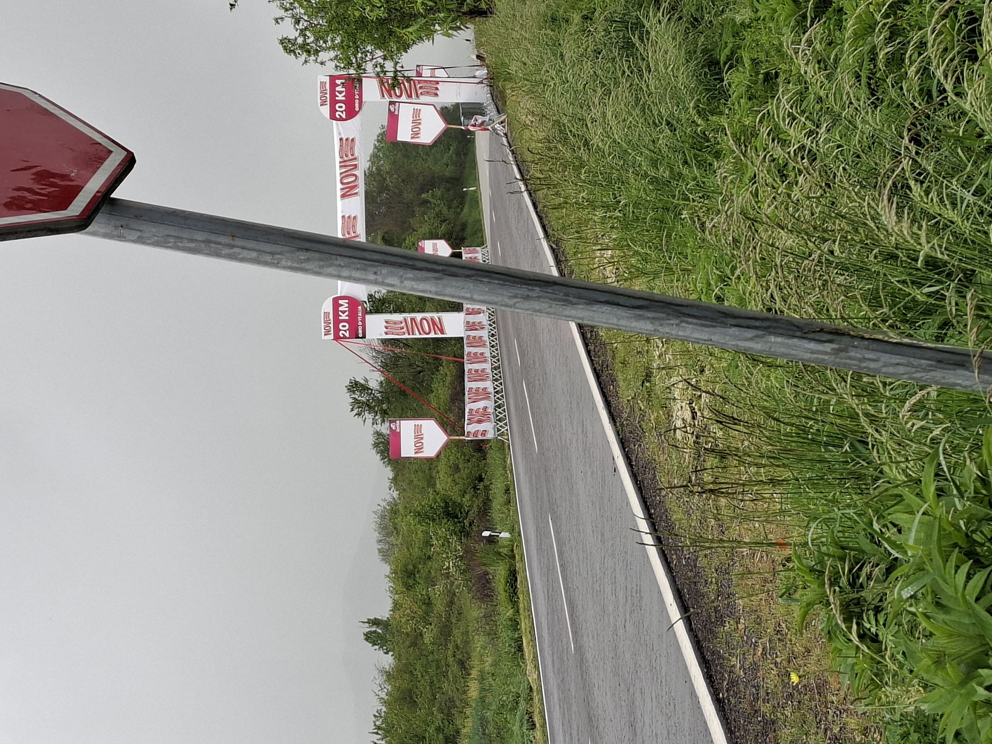 The image shows a wet road in Dragijevo, Bulgaria, lined with green vegetation on one side. The road is marked with a white line along the edge and a dashed white line down the center. Roadside signs and banners, indicating "NOVI" and "20 KM GIRO D'ITALIA", are placed along the road. A stop sign and its metal pole are visible in the foreground. The sky appears overcast.