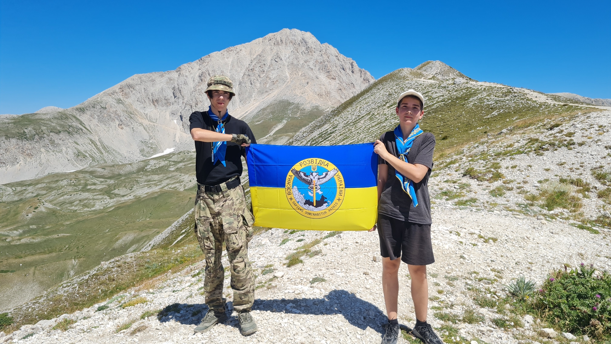 Two young males, appearing to be teenagers, stand on a rocky, mountainous terrain under a clear blue sky. The male on the left wears a camouflage hat, a black t-shirt, camouflage pants, and a blue scarf. He has his arms crossed and is looking towards the camera. The male on the right wears a white cap, a dark grey t-shirt, dark shorts, and a blue scarf. He is also looking towards the camera. Between them, they hold a Ukrainian flag, which is horizontally divided into blue and yellow sections, with a circular emblem in the center. The emblem features an eagle with outstretched wings, holding a sword and a shield, and text in Ukrainian and Latin.
The background consists of rugged, light-colored mountains with sparse green vegetation in the lower valleys. The terrain is characterized by gravel and rocks. The overall environment suggests an elevated, possibly remote, natural setting. The location is identified as the Gran Sasso mountain range, near L'Aquila, Italy.