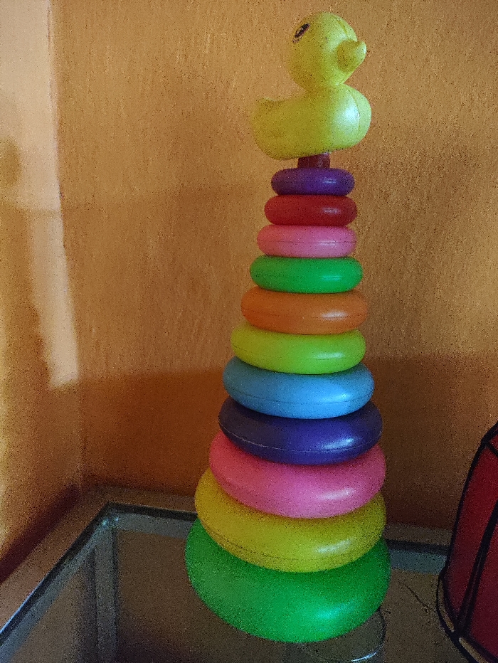 The image displays a vibrant stacking ring toy, topped with a bright yellow rubber duck, set indoors in Selagai Lingga, Indonesia. The toy is assembled into a pyramid shape on a reflective, dark surface, possibly a glass tabletop, which shows the toy's reflection. The rings are arranged in decreasing size from bottom to top, showcasing a variety of colors including green, yellow, pink, dark purple, blue, light green, orange, red, and another dark purple ring, culminating in the yellow duck figure. The duck has small black eyes and a reddish-orange beak.
The background consists of a textured, warm orange-brown wall, suggesting a domestic setting. No activity is depicted; the image is a still life focusing on the children's toy. The lighting appears to be ambient indoor light. A dark, possibly red object with a metal frame is partially visible on the right side of the frame. There is no visible text in the image.