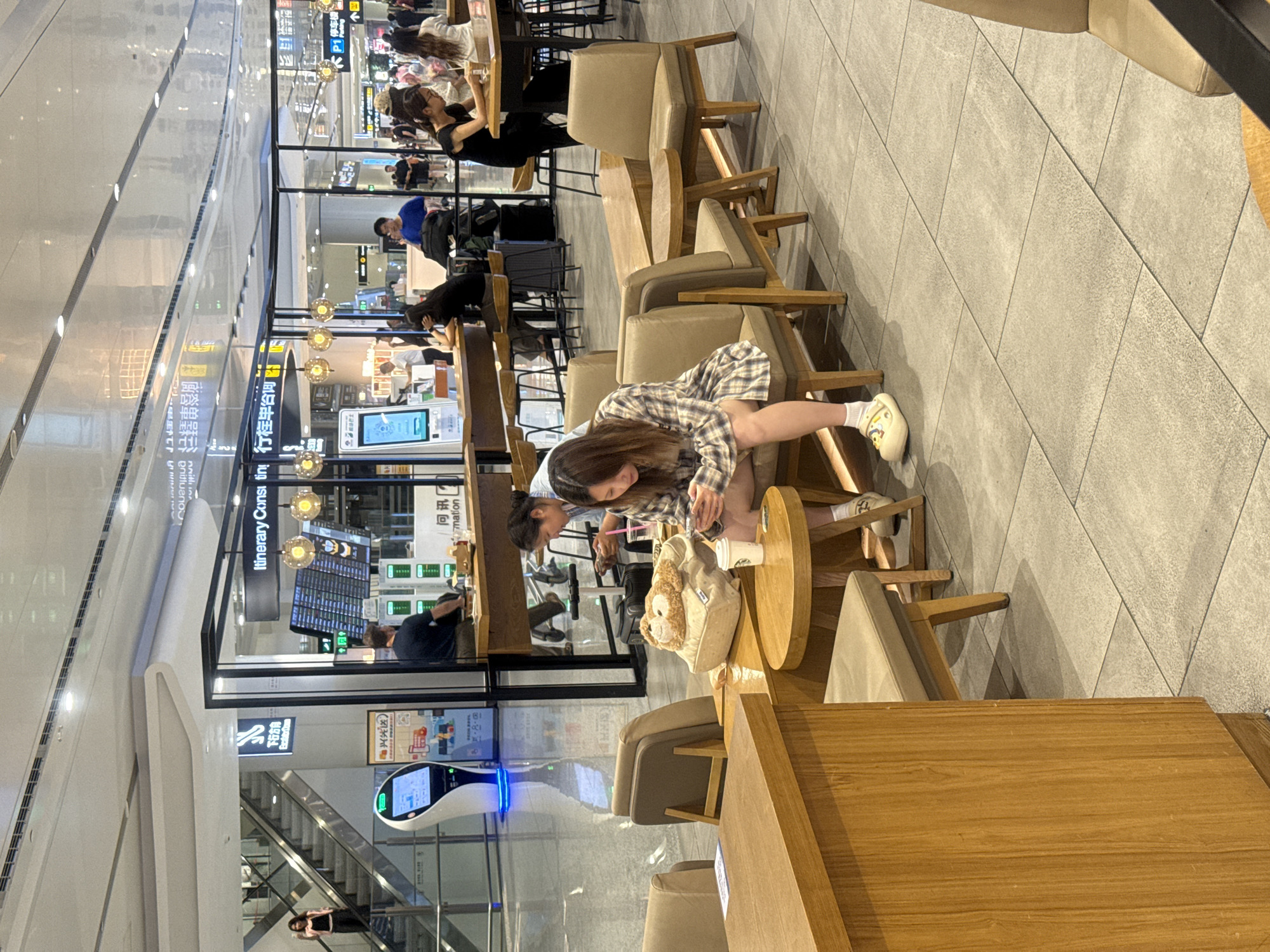 This is a candid, eye-level medium shot taken in a modern indoor public space, likely an airport or large transit hub, with a sleek, reflective ceiling and tiled flooring. The scene is set in Guangyang District, China. In the foreground, a young woman with long, dark hair, wearing a plaid shirt and shorts, is seated at a small round wooden table. She is looking down at her phone, holding a white cup in her right hand, and a plush toy sits on a cushion next to her. She is wearing light-colored clogs with a yellow motif. Adjacent to her are two tan armchairs and a larger wooden table. Further back, a man in a blue shirt is standing at a counter with an information display. To his left, there is a digital kiosk with a blue illuminated base. Behind the counter, several people are seated at tables. In the background, escalators lead up to a higher level, and various signs, some in Chinese characters, are visible. The overall atmosphere appears to be one of quiet activity and public waiting.